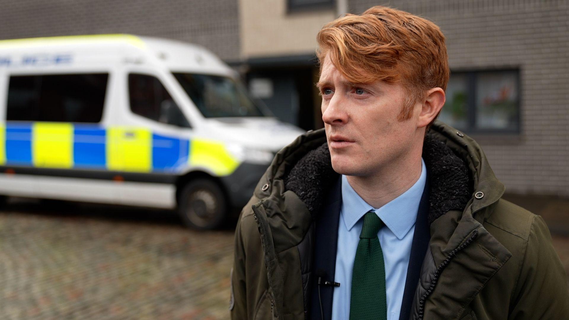 Det Insp Alan Swanston is a red-haired man with a blue short, green tie and green coat. He is standing in front of a police van outside a modern building.