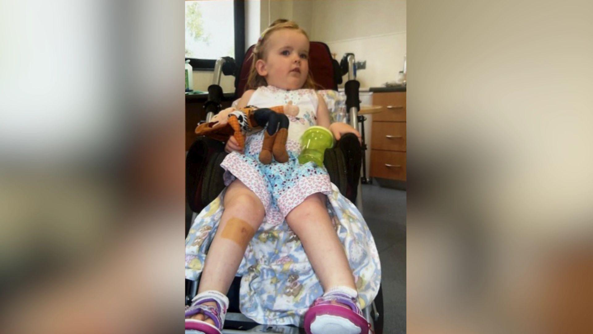 Emily in a wheelchair as a young child. She is wearing a colourful patterned dress and holding a Woody from Toy Story toy. She has short blonde hair. 