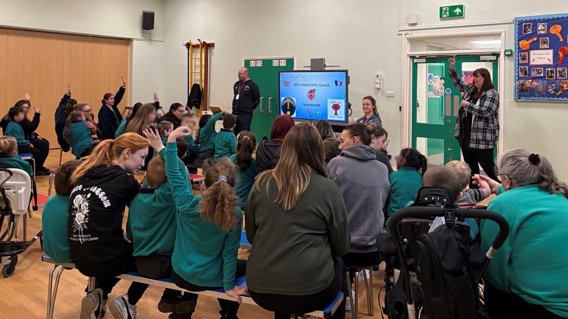 The view at the back of a school assembly. Pupils are wearing a teal blue coloured uniform. Maisie Summers-Newton is addressing the pupils at the front, next to a TV screen showing her Paralympic achievements.