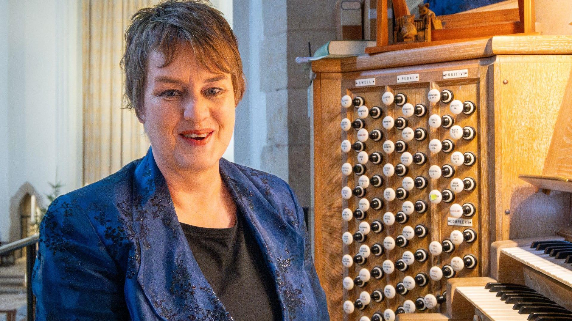 A woman with short hair wearing a blue suit jacked with a black pattern on it. She is sat at an organ, with rows of keys and handles visible to her side.