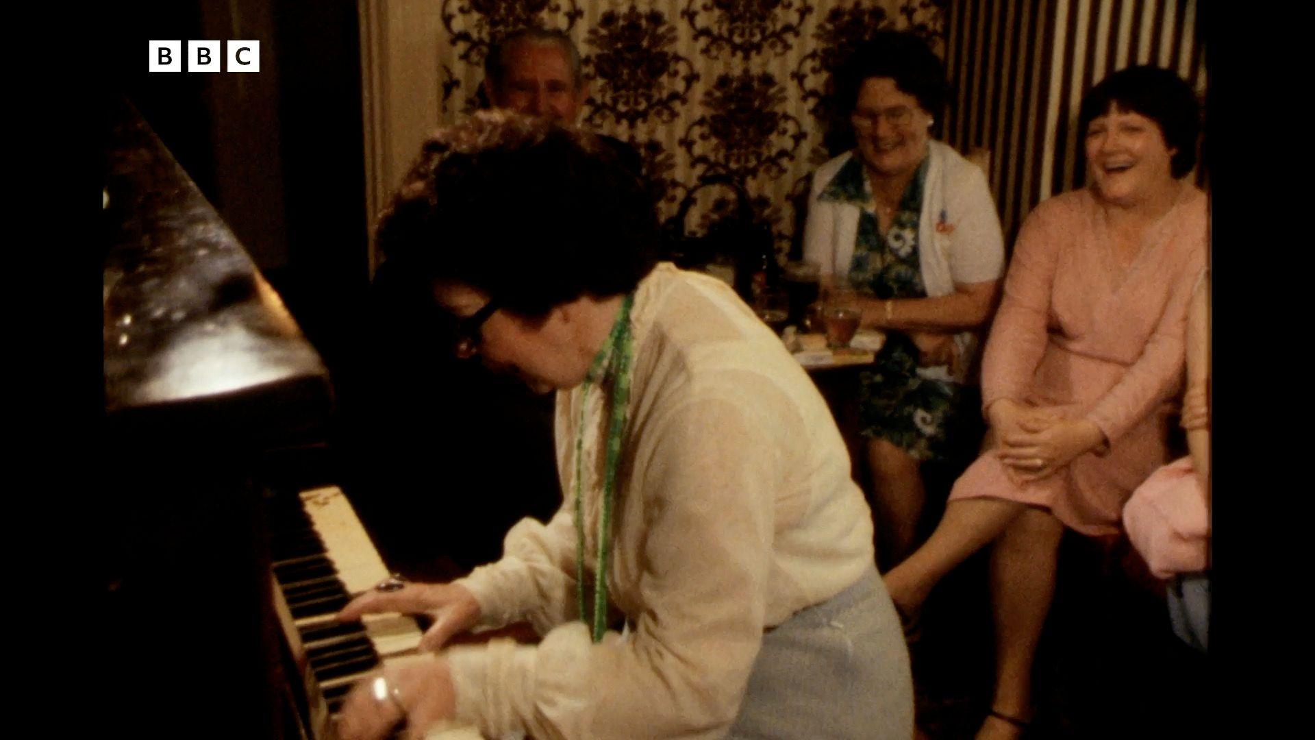 Peggy playing the piano in a pub. Three people are sat behind her watching and laughing.