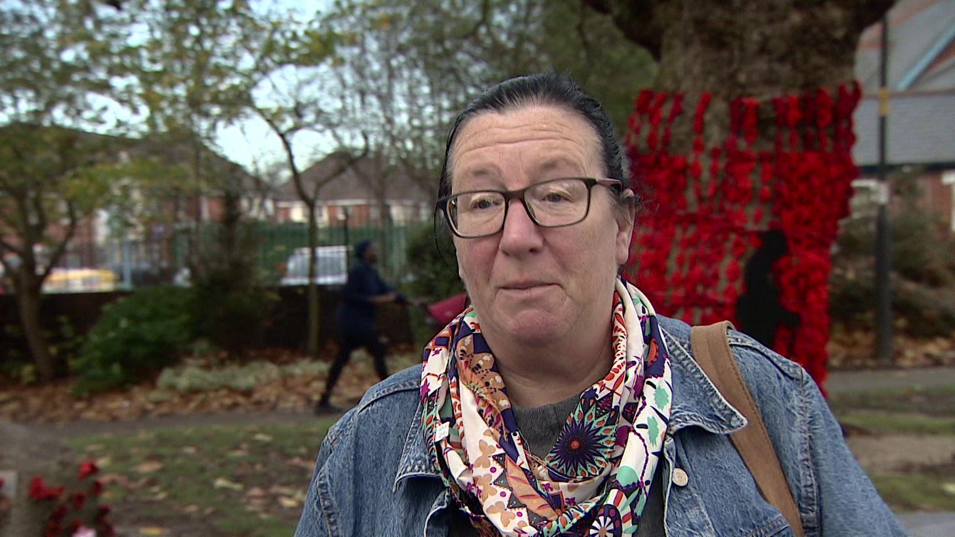 A woman with glasses is wearing a neck scarf and jacket. She is standing in park in front of a tree that has been decorated with remembrance poppies.