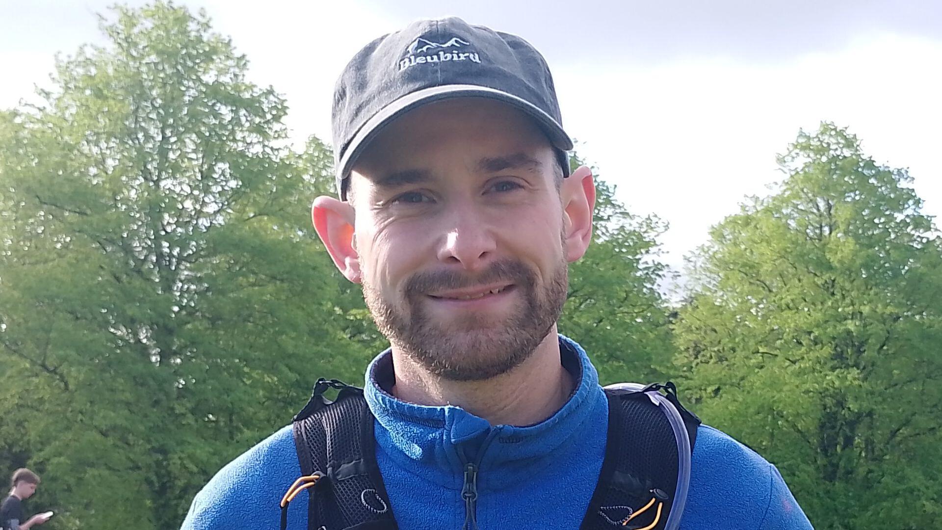 Michael wearing a black cap, blue half zip fleece and black running fence smiles at the camera. He has a dark beard and moustache.