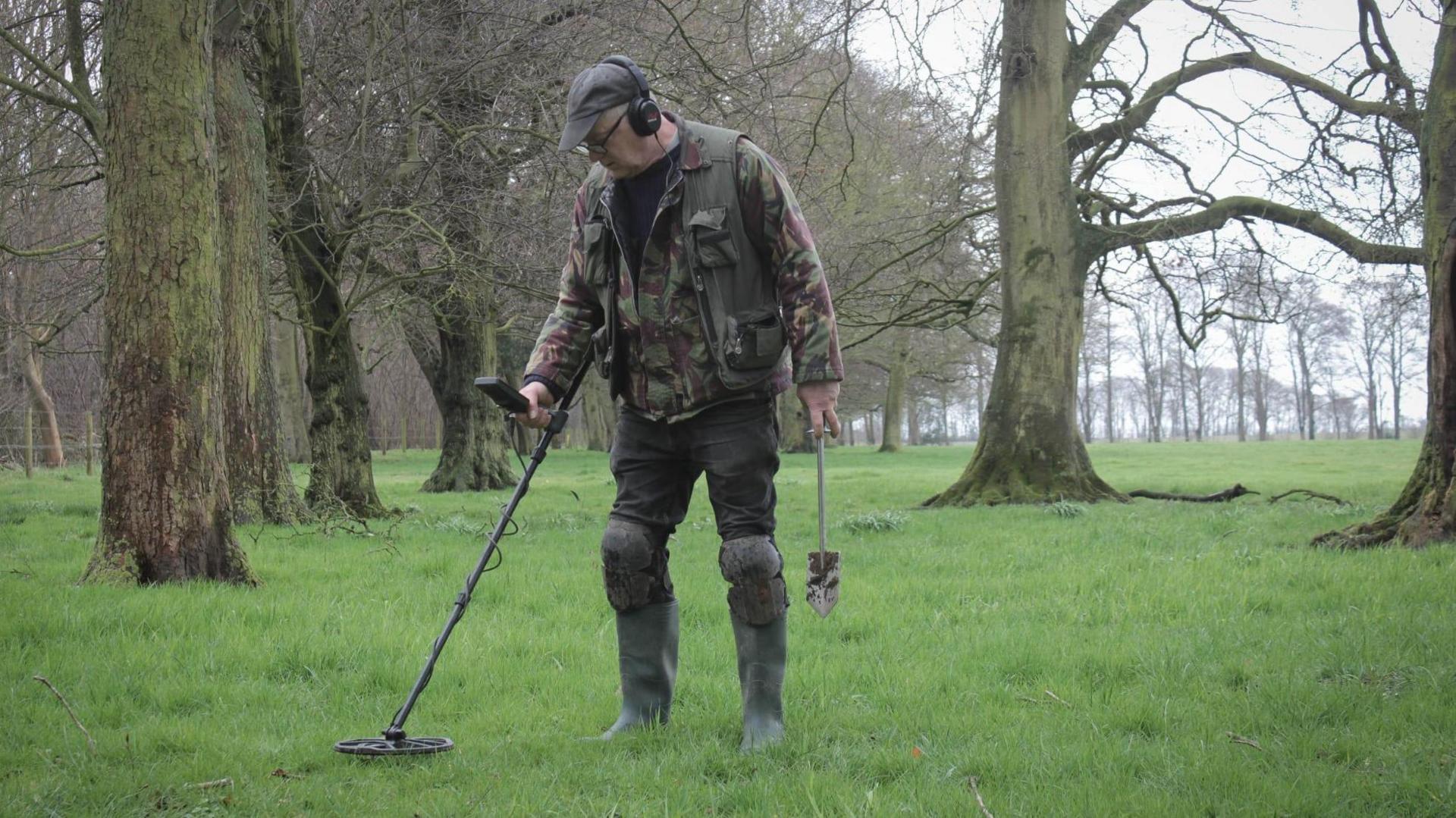 David Stuckey is standing in parkland which has lots of green grass and large trees. He is holding a metal detector and a small spade for digging. He is wearing a camouflage jacket and knee pads over his trousers which are tucked into green boots