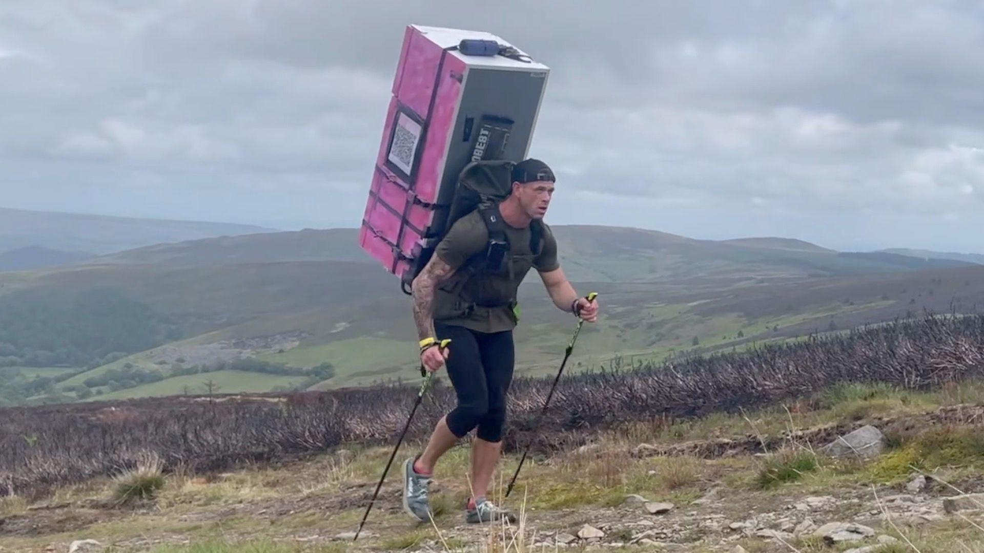 Matt Jones walking on the hills with pink fridge freezer on his back in leggings and khaki green t-shirt