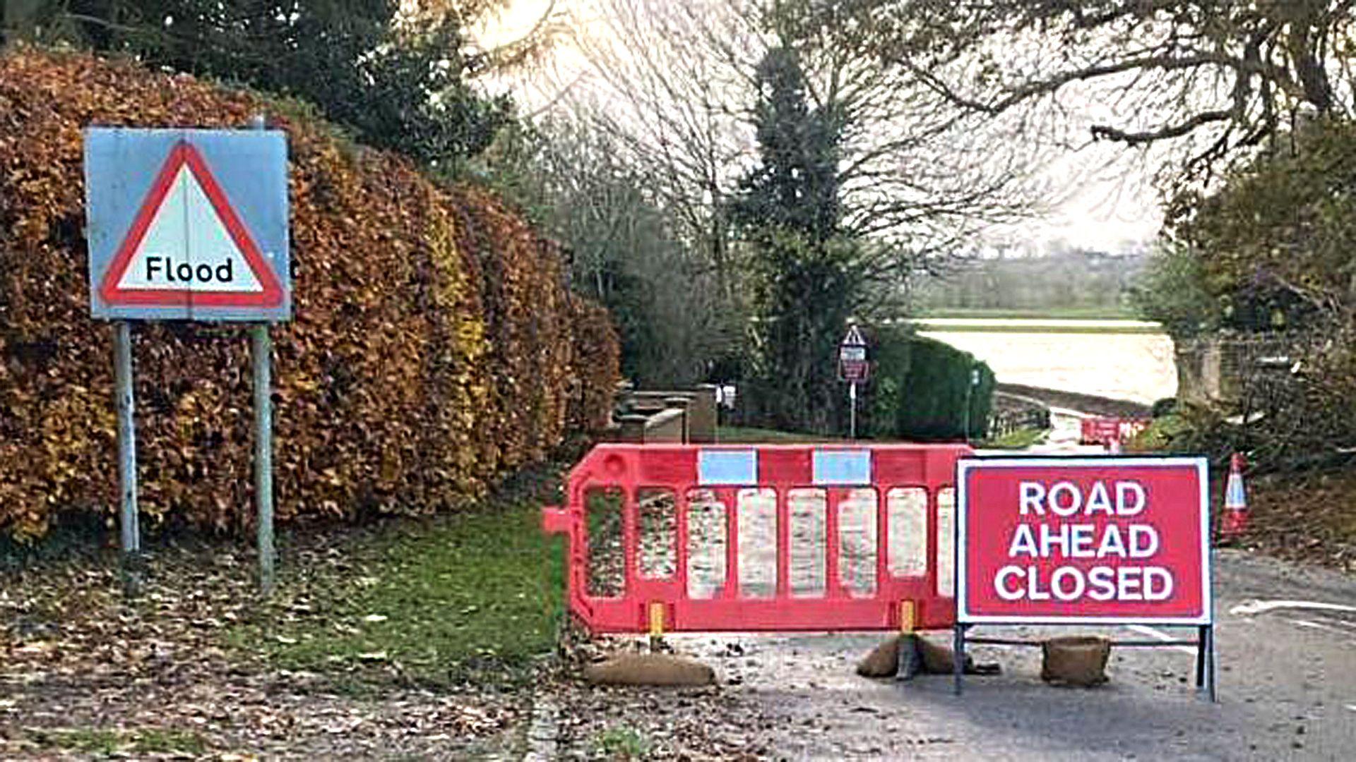 Radwell road closure showing a Road Ahead Closed sign and a large red barrier. You can see a flooded river in the background, flood sign, trees and bushes. 