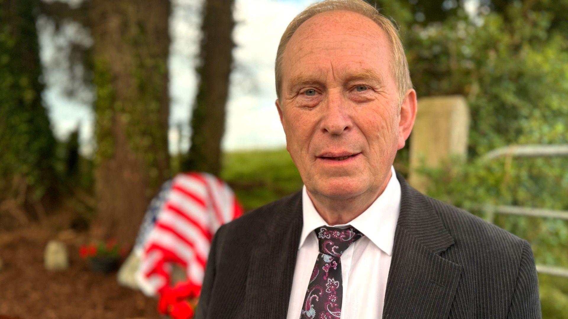 An older man with fair, receding hair. He is wearing a dark pinstripe suit with a white shirt and a purple paisley tie. He is standing in front of a field gate. Also behind him, out of focus, are some trees, and the memorial stone which is draped in the American flag.