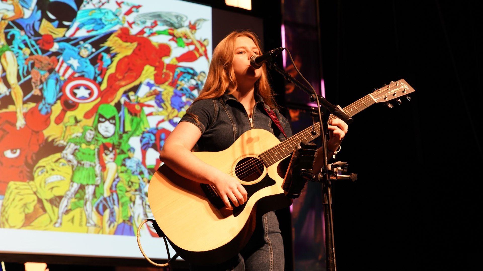 Liv Jones, a young woman with long hair, stands singing and playing an acoustic guitar on a stage.