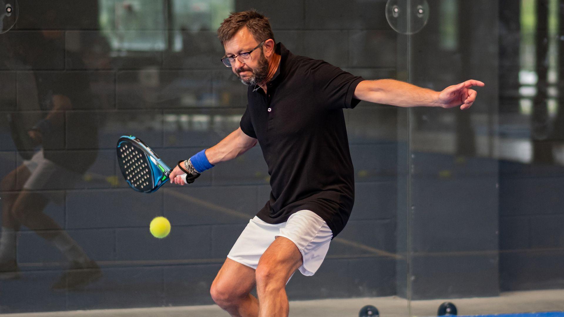 A man with a stern look about to hit a forehand with a padel racket, at an indoor centre. He has black hair, a black and grey beard, glasses and a necklace. He is wearing a black top and white shorts.