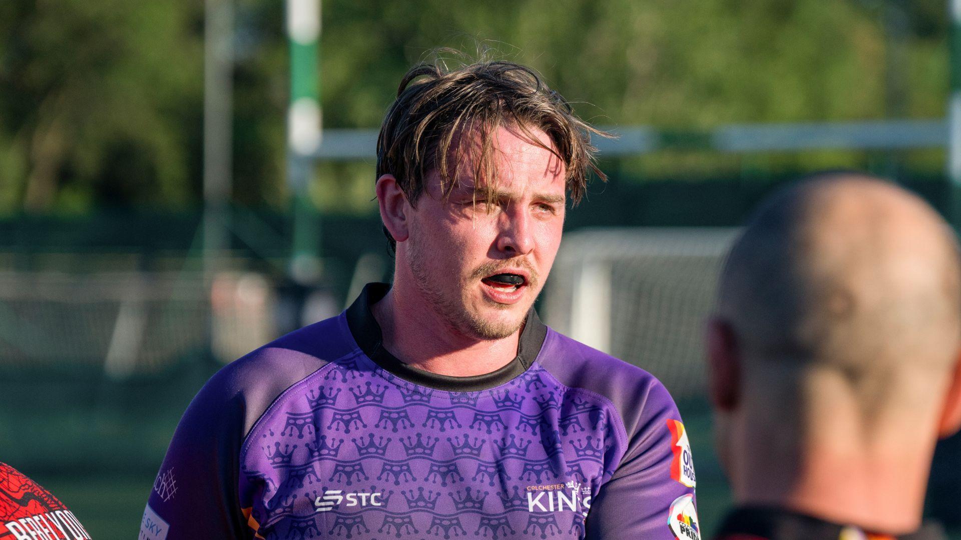 Tom Crudgington during a rugby game. He is wearing a blue rugby shirt and a gumshield and his brown hair is ruffled. He has a short brown beard and a moustache. The back of another player's head is also in the shot.