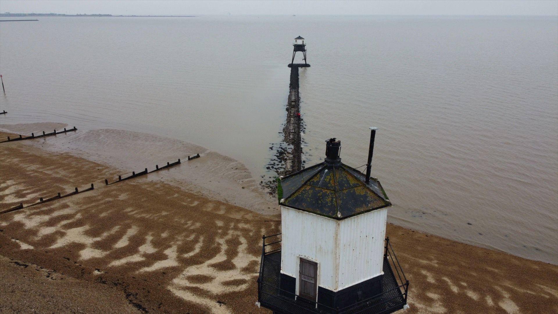 The leading lights, a pair of Victorian lighthouses, standing on the beach at Dovercourt. One is in the foreground on the beach, the other is out at sea, joined by a causeway. 