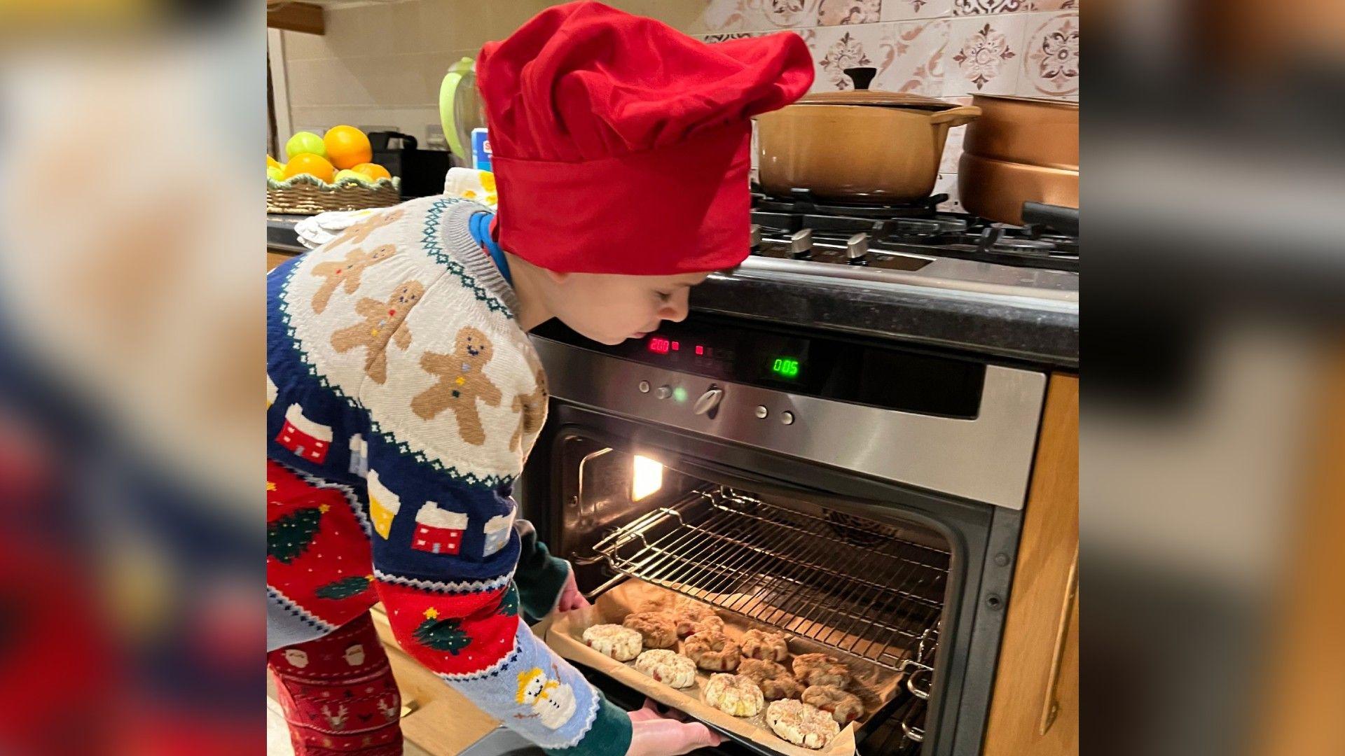 Joshie Harris pictured putting a rack of pies in the oven in his kitchen. He is wearing a red hat and a multi-coloured jumper with gingerbread men on and red trousers with Santa on.
 