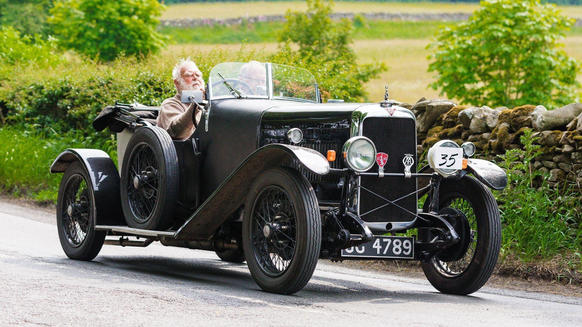 A very old open-top car is driven along a country lane by a man with a white beard.