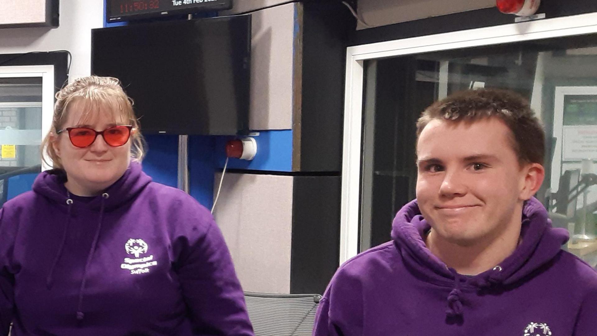 Donna Wyatt and Hamish Crawford are pictured next to each other inside a radio studio. They both wear purple hoodies and are smiling.