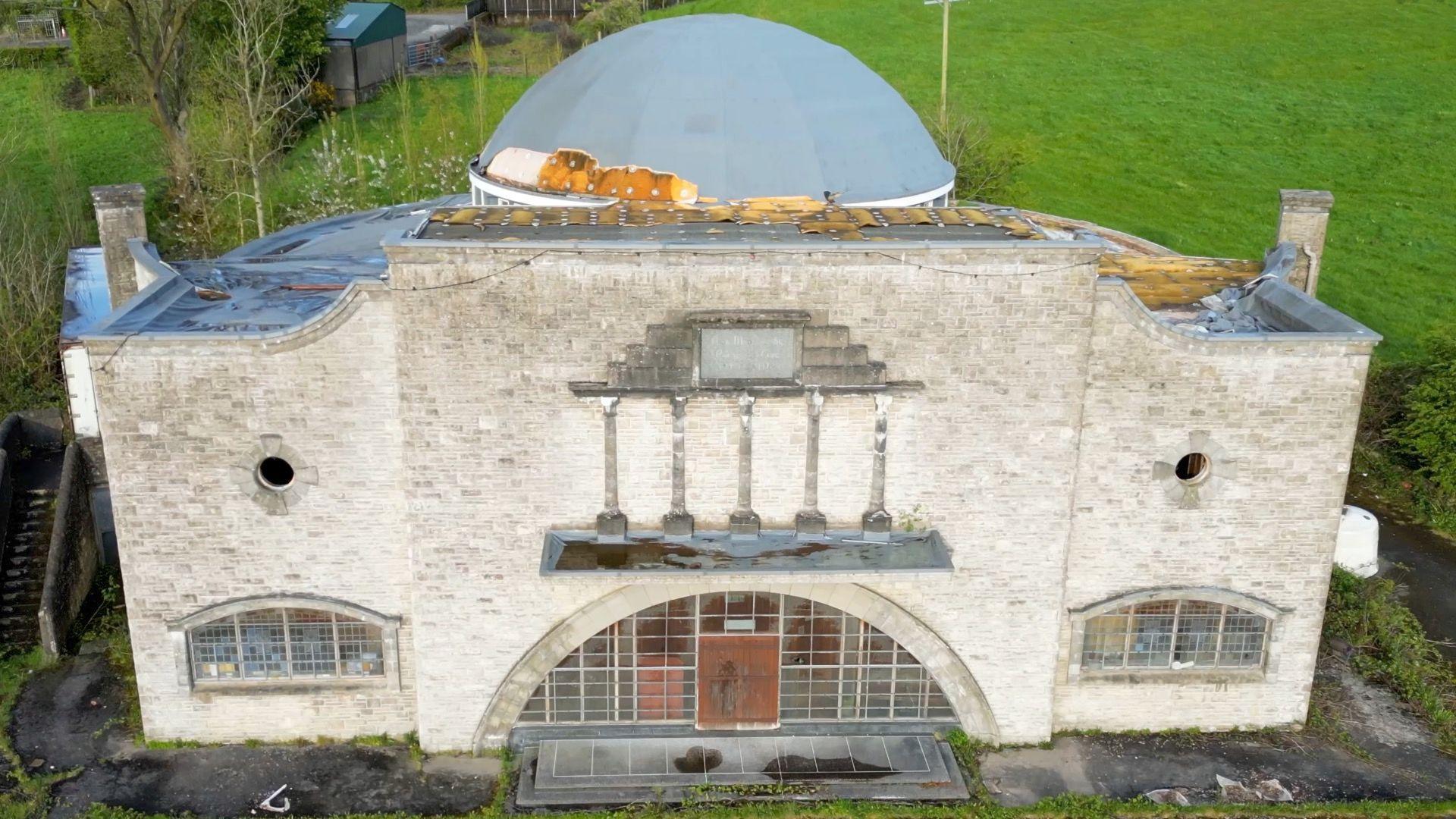 Edendork Hall taken before its demolition with a light brick exterior and curved large windows, at the top of the building is an impressive large dome roof which is light grey with specks of gold (it would have been gold all over before deterioration. It stands on a plot of land with green grass behind it. 