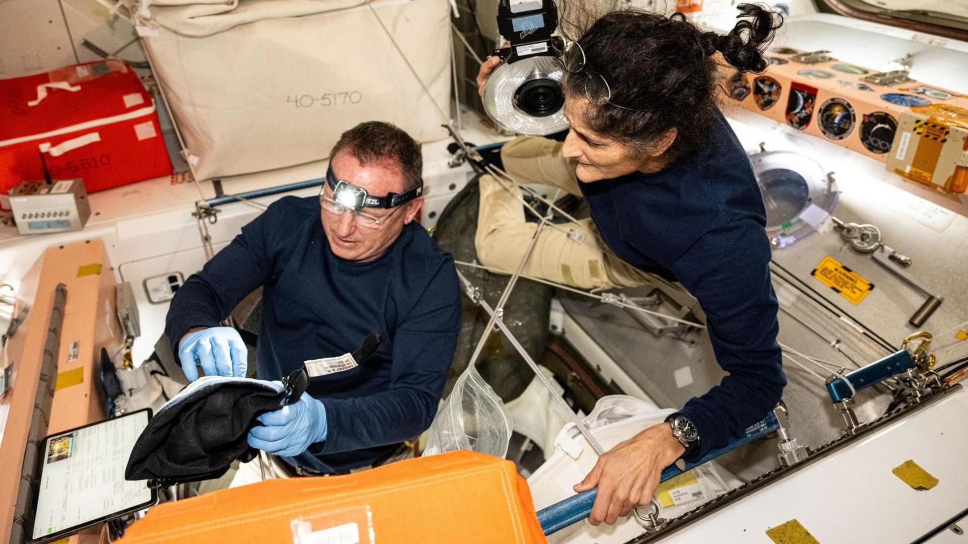 Butch and Suni carry out an experiment - Butch wears a headlight and blue surgical gloves. He is sitting in a type of pit and appears to be looking inside a black bag. Suni hangs just over him, looking at what he is doing. 