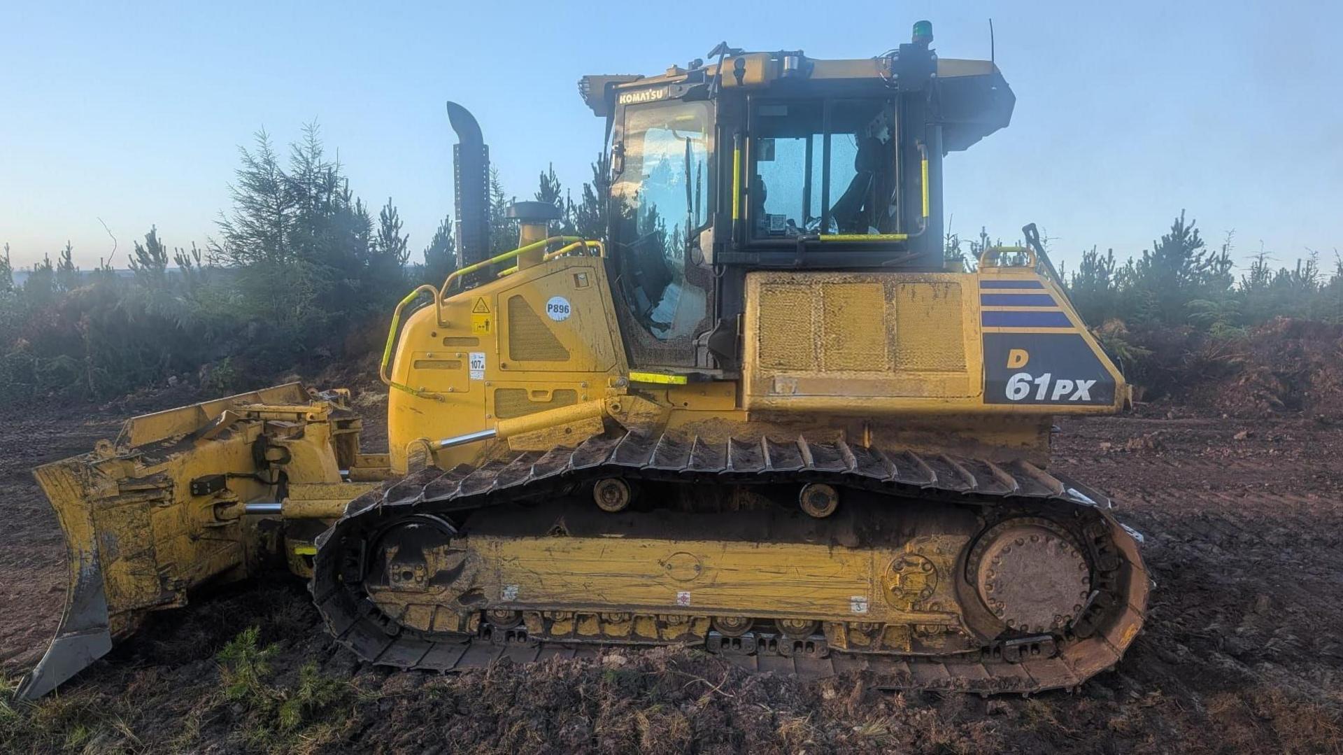 A yellow bulldozer driving over churned up soil with trees behind it
