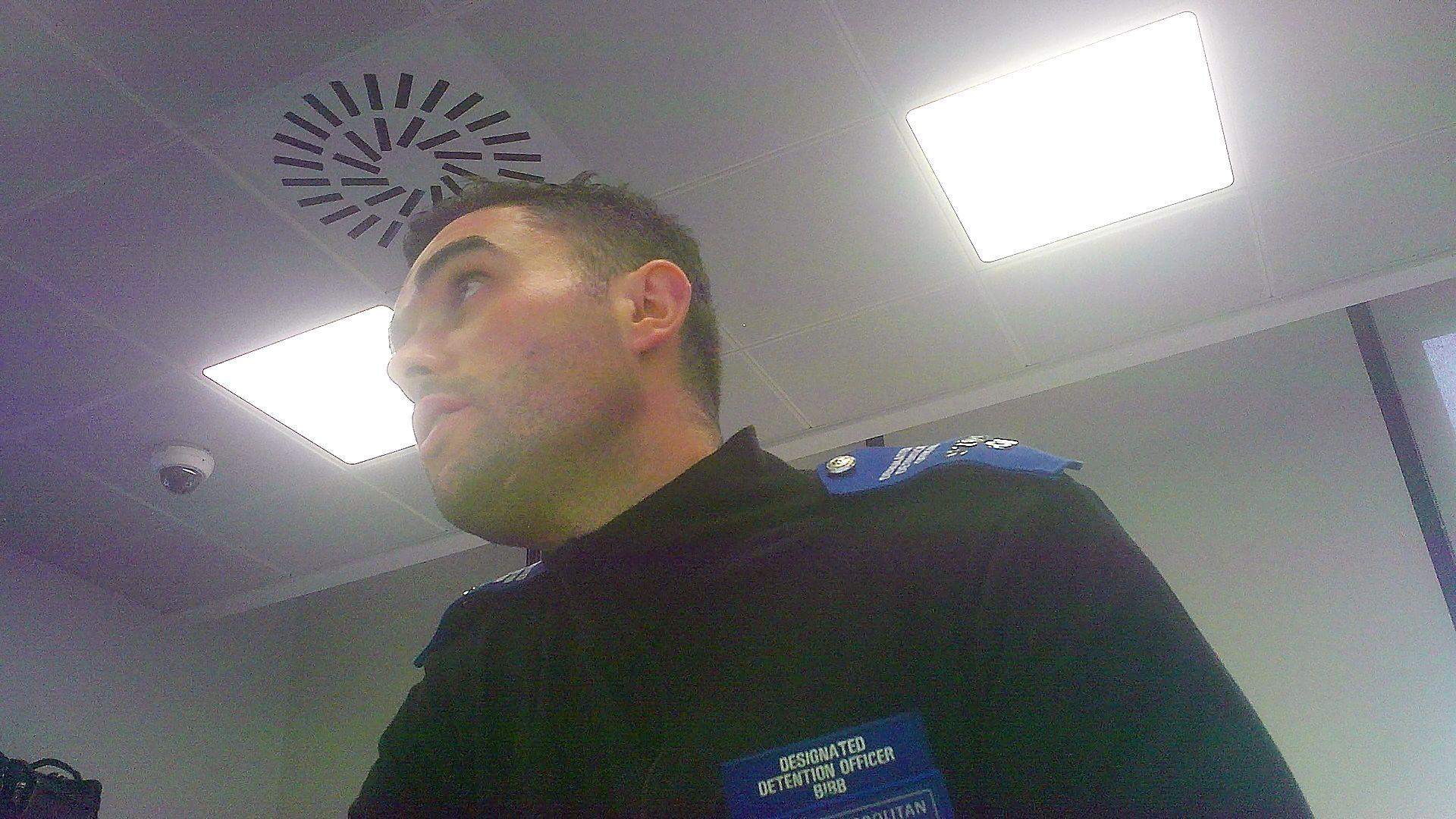 A hidden camera image of Rory Bibb in his uniform, inside Charing Cross police station. He is filmed from below, and on the ceiling above him can be seen a dark glass dome, which appears to be a CCTV camera