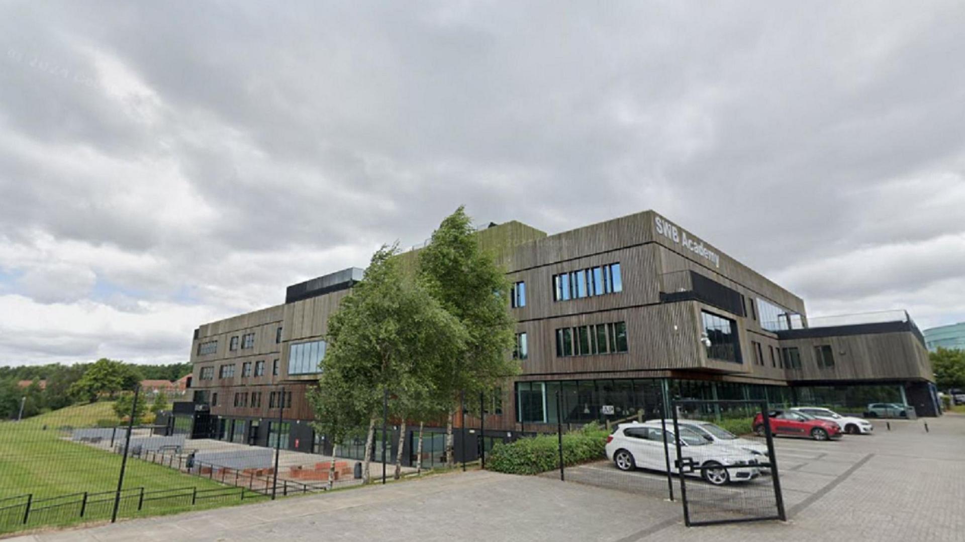 A large school building with SWB Academy in large white letters on the side of the building. There are trees and a playing field as well as cars parked in a car park behind mesh fencing.