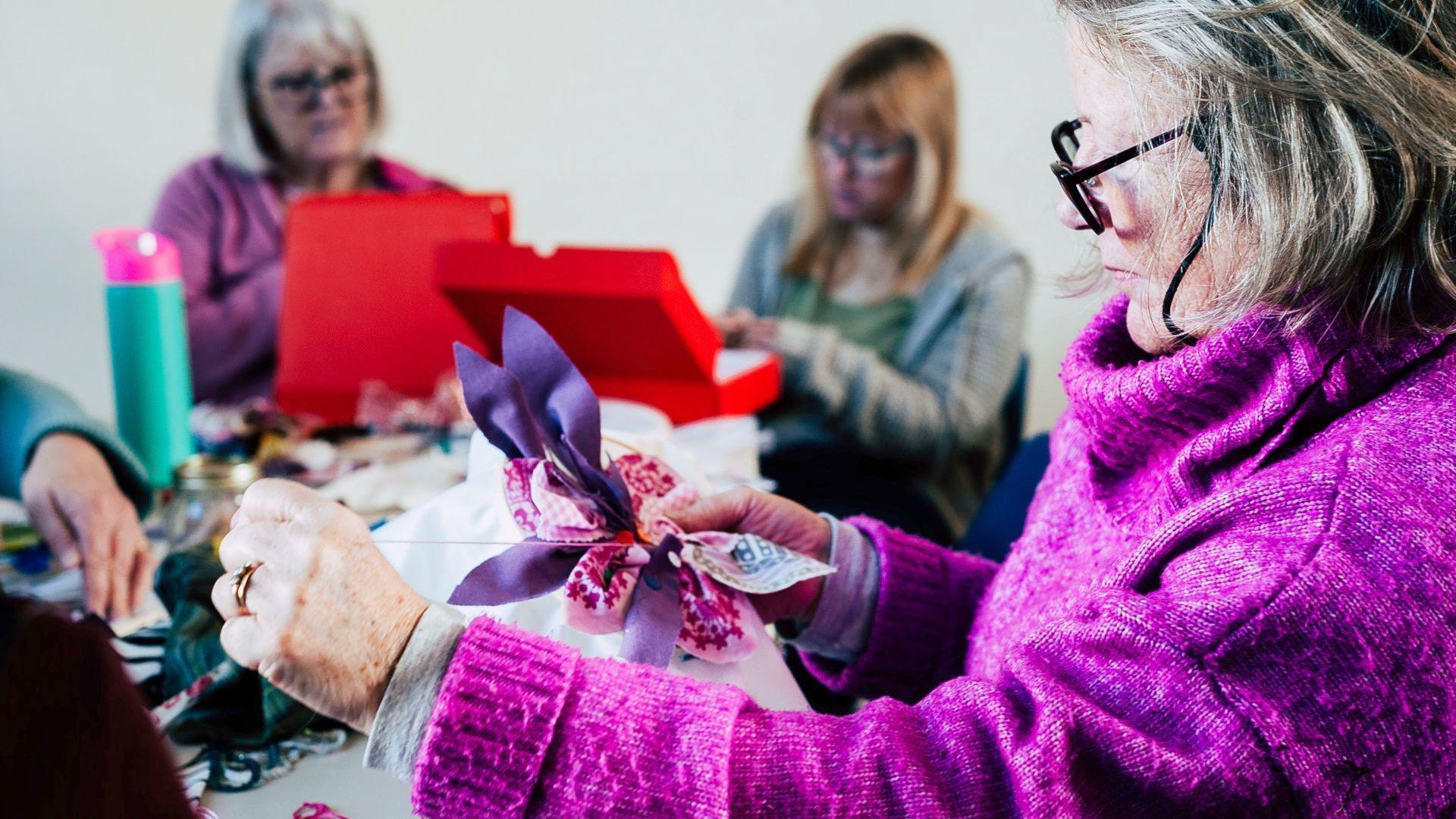A close up of a woman sewing. She has grey/blonde short hair and wears black framed glasses and a bright purple thick woollen turtleneck jumper. She is sewing purple ribbong and pink floral fabric onto a piece of white calico material. In the background, which is slightly blurred, two other women sit sewing.