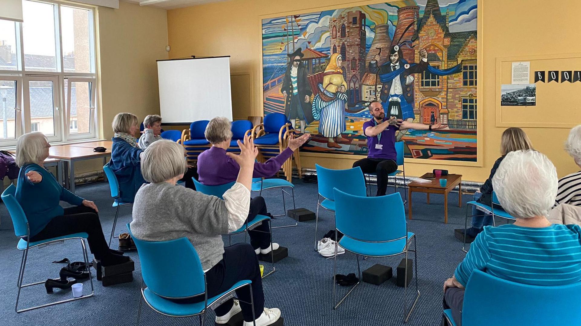 A group of women sitting on blue chairs facing away from the camera. A male instructor sits facing the women with his arm out stretched.