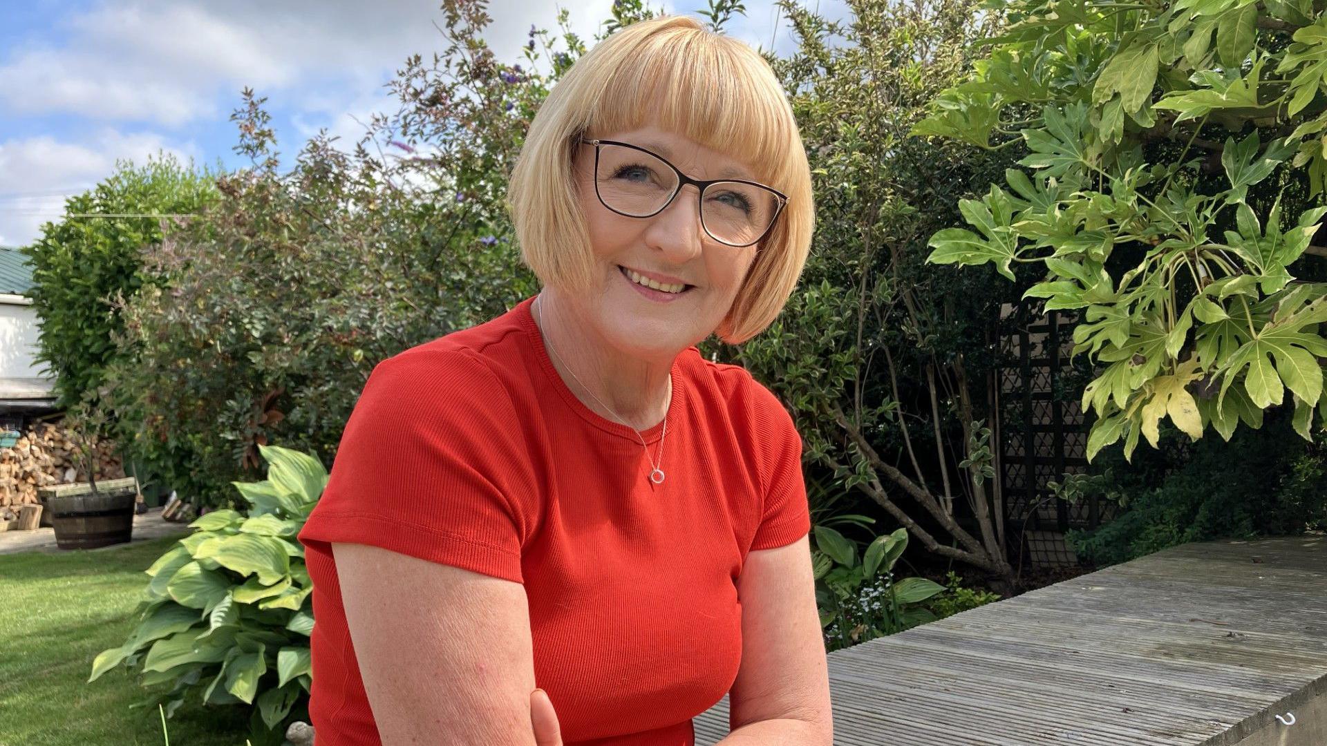 A woman sits on a bridge over a garden pond, facing the camera. She's got chin length blonde hair, and is wearing a bright orange t shirt. She's wearing black rimmed glasses, and is smiling. Trees and plants are visible in the background.