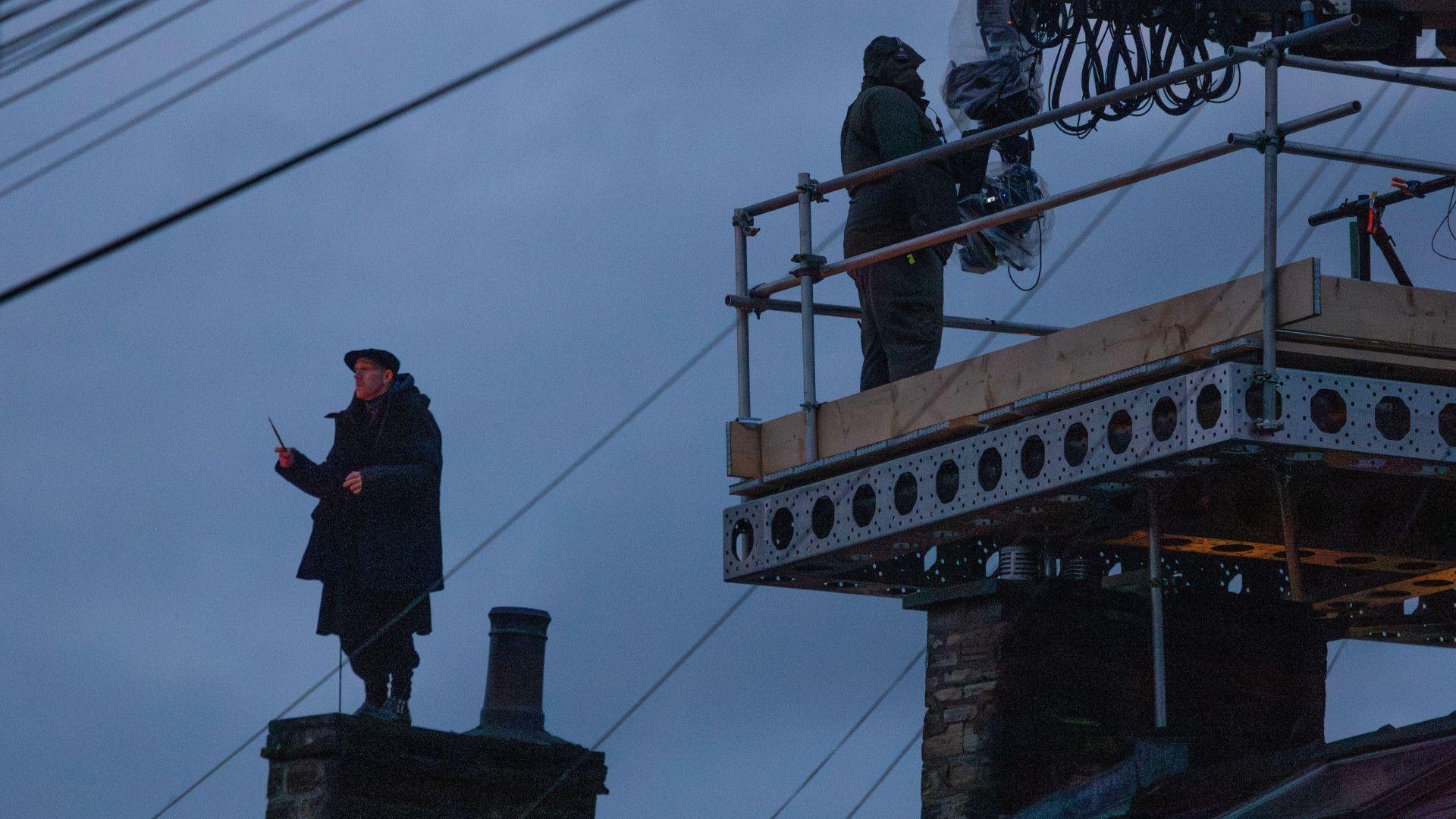 A man dressed in dark clothing stands on top of a chimney in the early evening light. He is wearing a flat cap and holding his right arm above his head with a twig-like wand in his grasp. A member of film crew can be seen standing on a platform in front of him