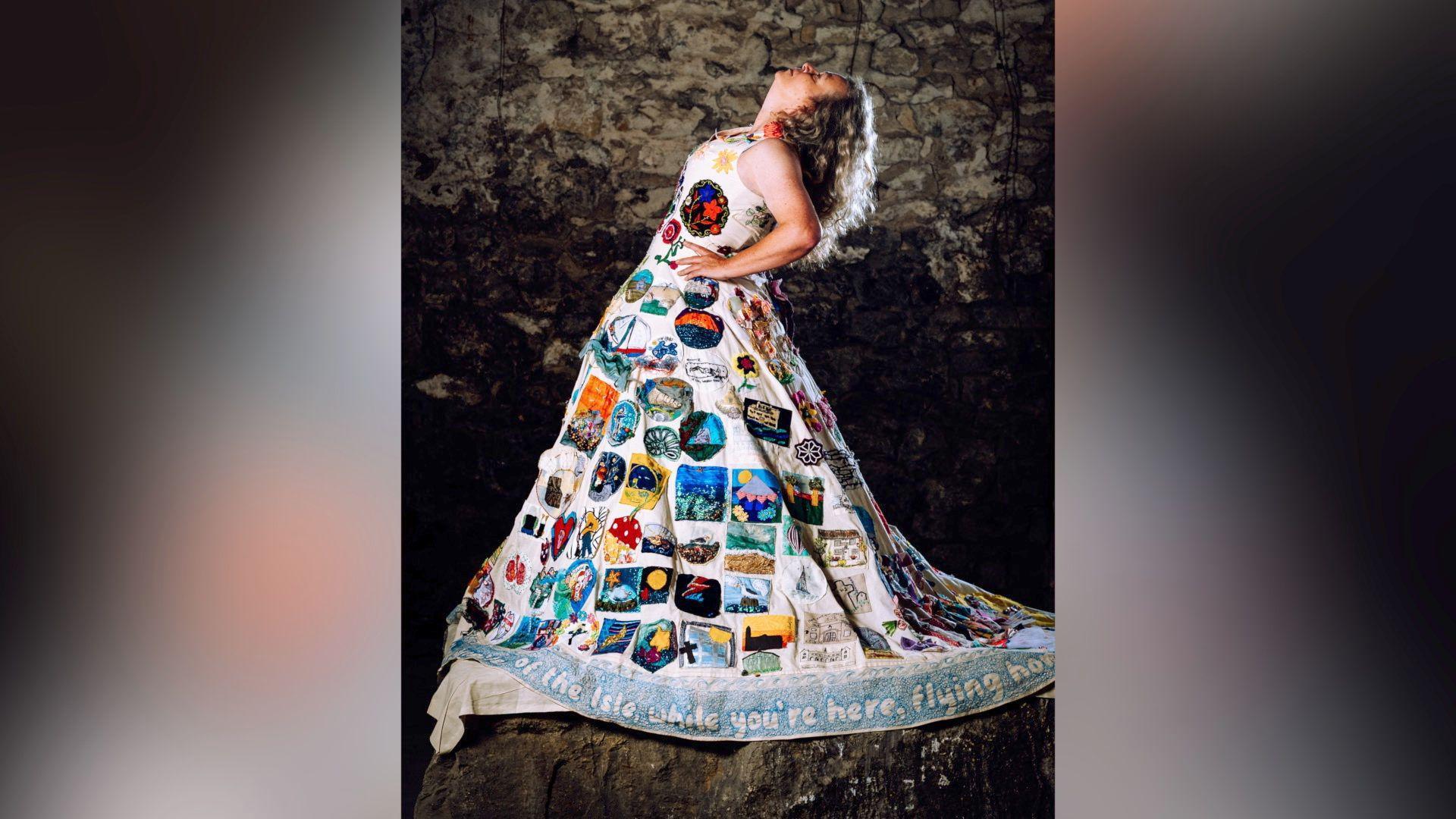 Sarah Partridge stands in what looks like a stone cave, on top of a large rock. She is facing sideways to the camera in a dramatic stance, her hands are on her hips and she is tipping her head back. Sarah has blonde curly hair. She is modelling the Portland Dress and from this angle the hemline can be seen with poetry written around the edging.