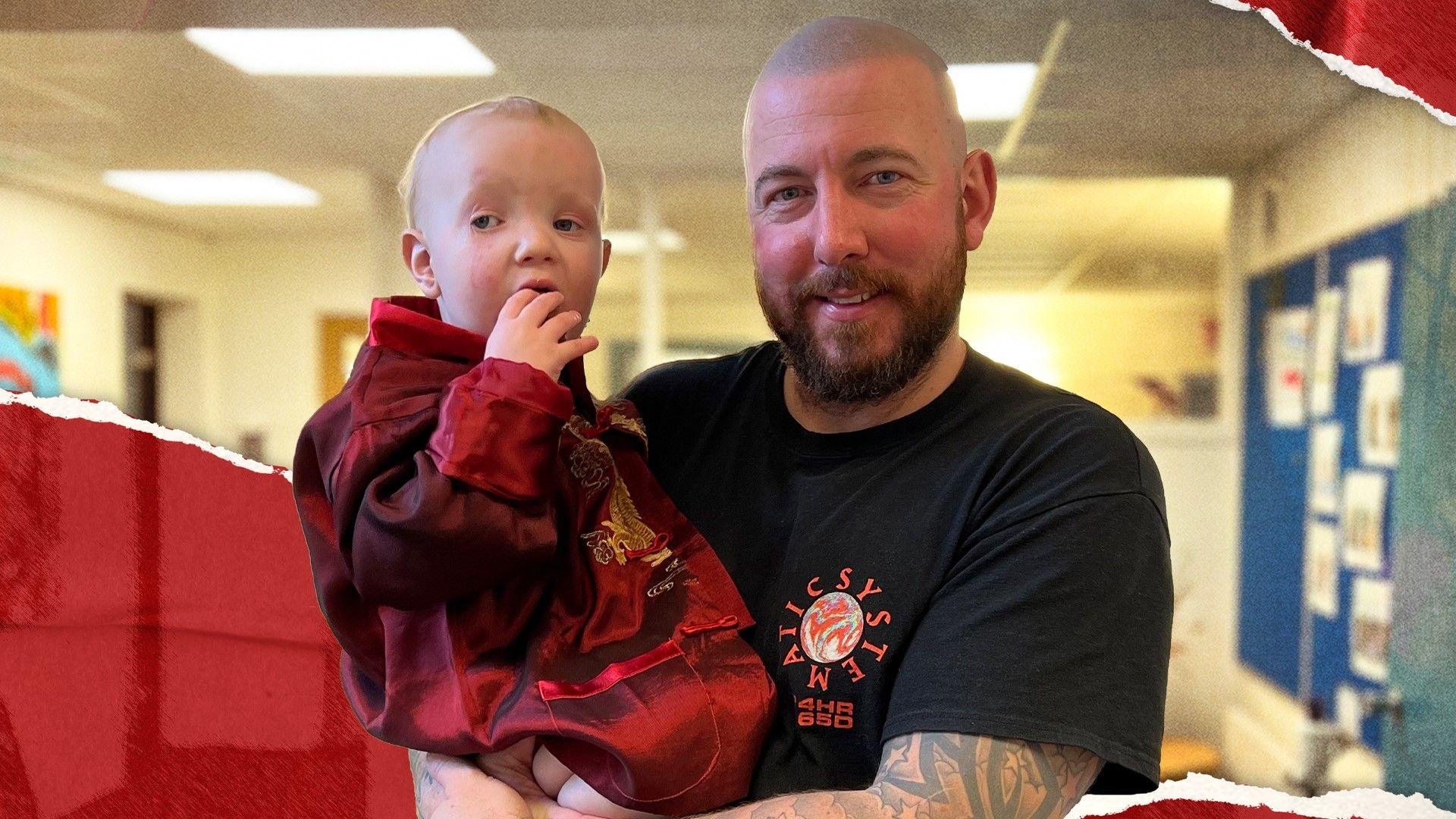 Andy Reay, a bald man with arm tattoos in a black t-shirt, holding his son who is in a red jersey, pictured in a classroom setting