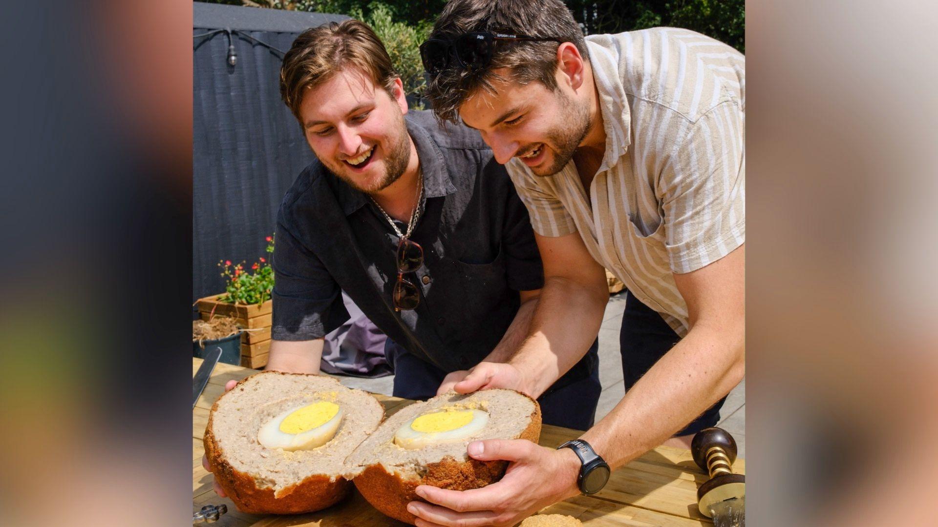 Oli and Phoenix are holding their massive scotch egg, which is on a wooden table, that's been sliced open to reveal a giant ostrich egg baked inside. They are looking down at their creation and smiling.