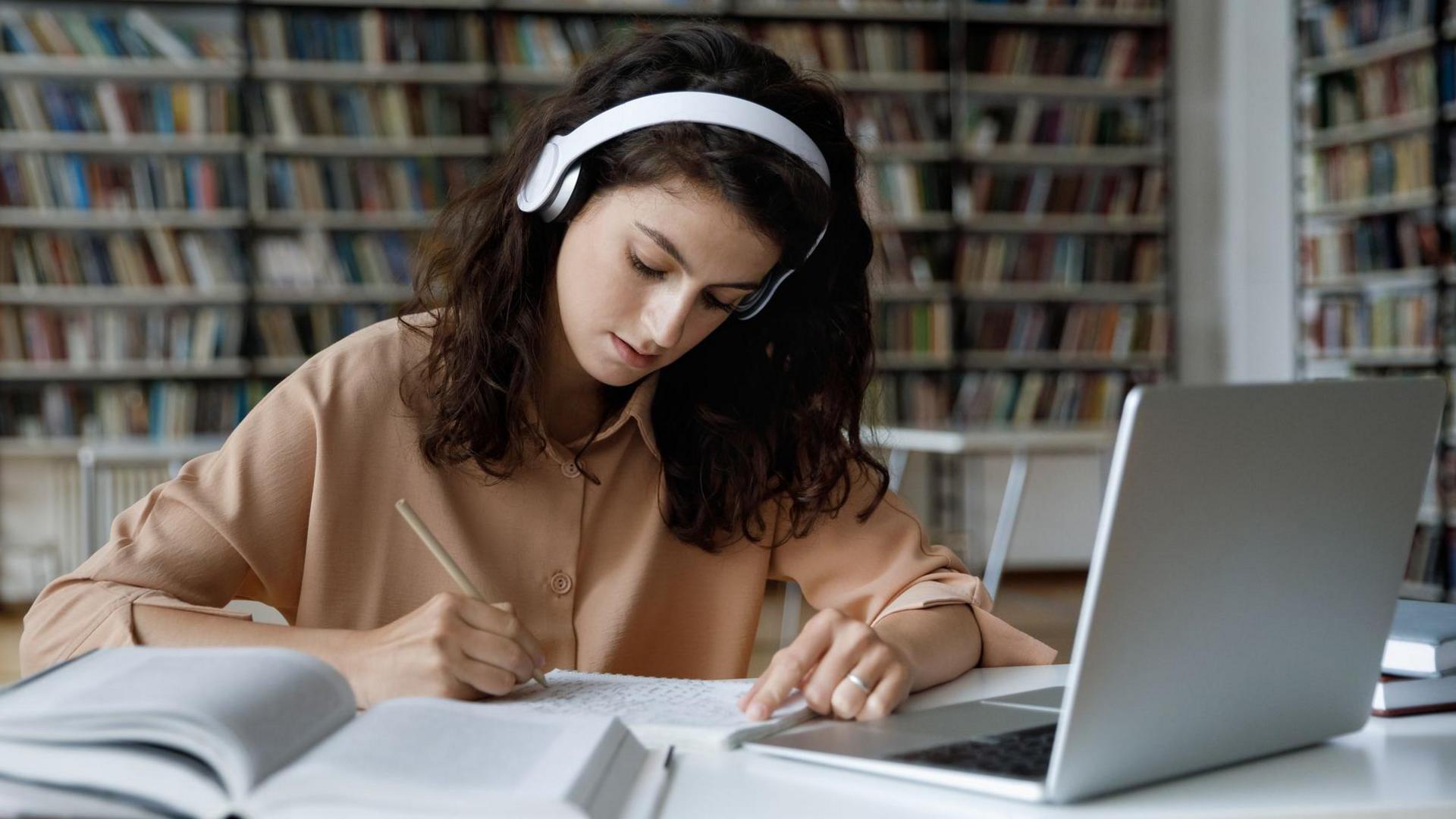 Woman with shoulder-length brown curly hair sits at a desk. She is looking at a piece of paper with a pen in her hand. There is a book and a laptop open in front of her. She is wearing a peach shirt. There are shelves of books in the background.