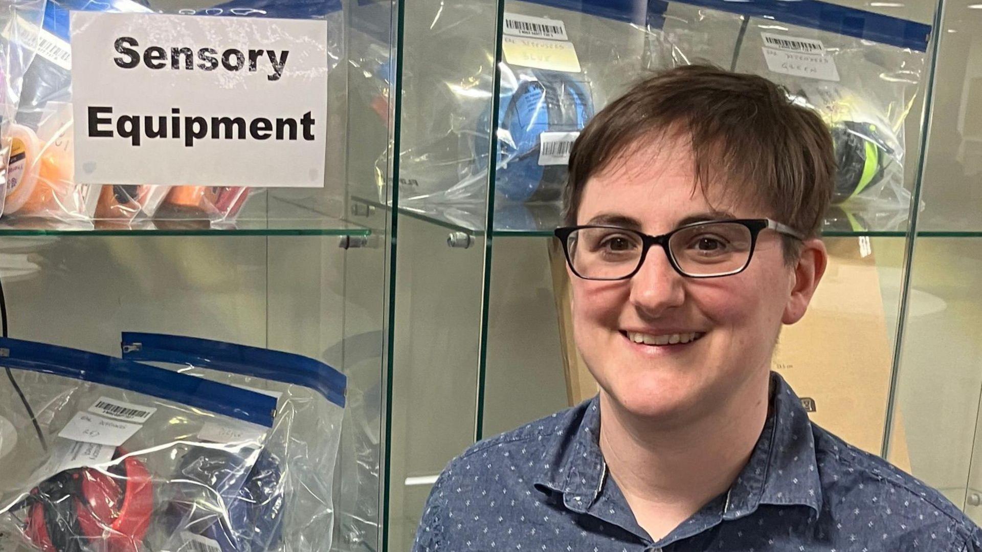 A person in a blue short-sleeved shirt stands smiling in front of a glass cabinet filled with noise cancelling headphones and fidget toys. A sign reading 'Sensory Equipment' is taped to the front of the cabinet.