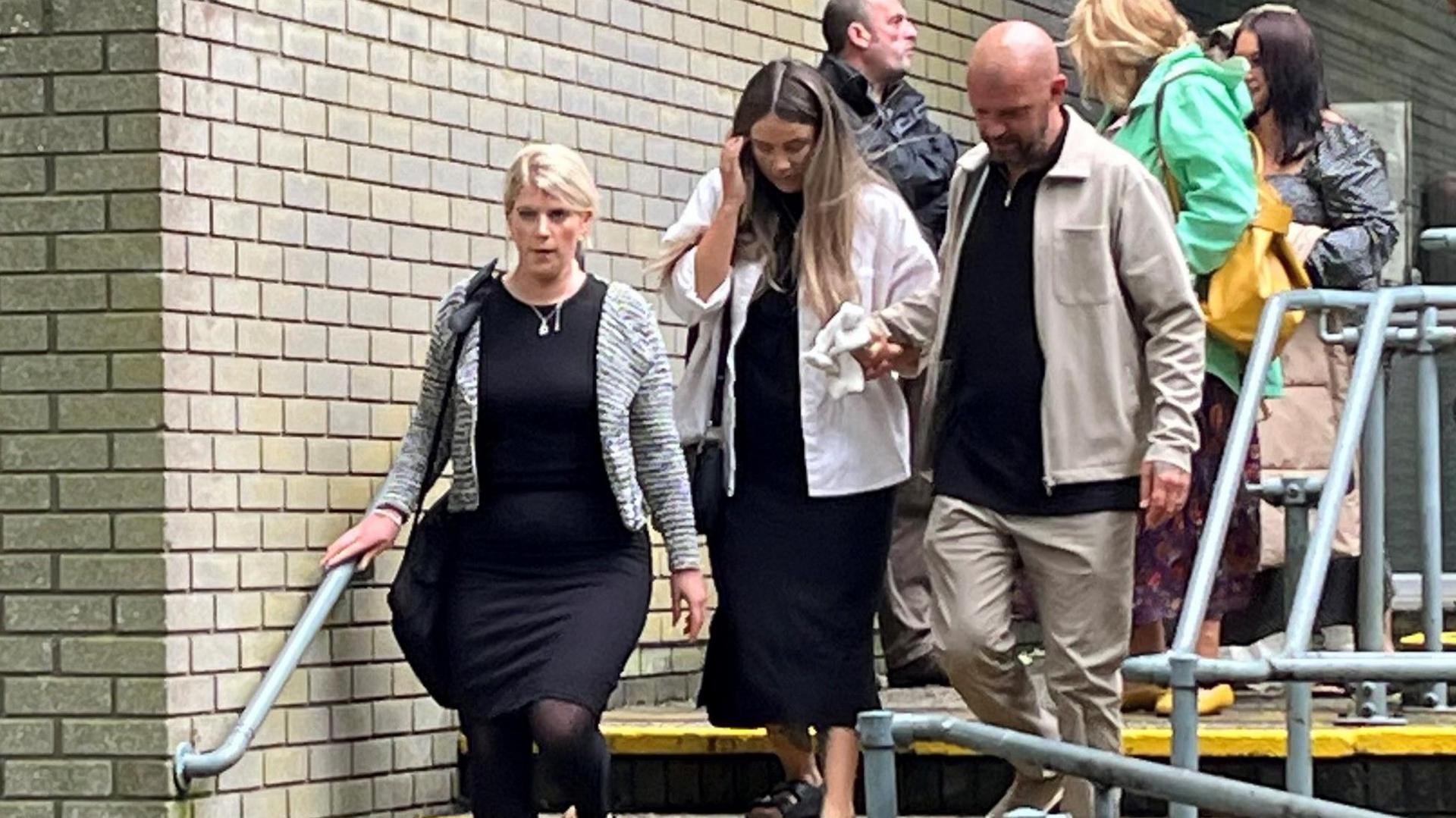 Mabli's family left court holding a soft toy. Two women wearing black dresses and light coloured cardigans walk down the stairs. One is holding a soft toy in her right hand which is also being held by a man who is bald and wearing beige trousers and jacket and a black top. Six other family members are visible walking behind them wearing coats. 