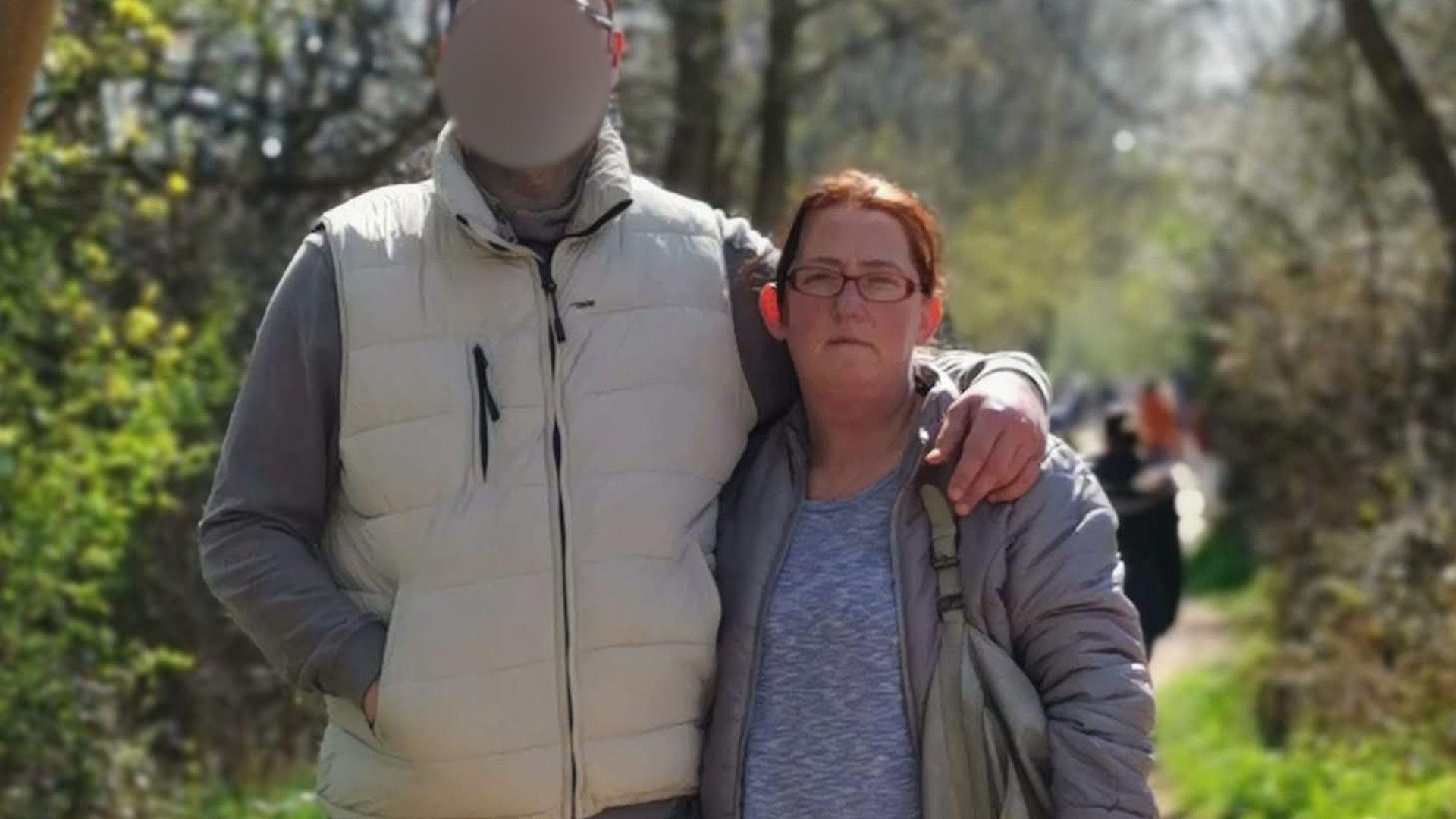 Marta Bednarczyk with short brown hair and brown glasses wearing a blue top and a grey coat with a grey bag, standing next to a man whose face has been blurred on a footpath lined by trees.