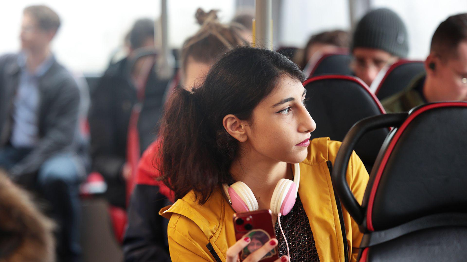A young woman wearing a yellow jacket and pink headphones is holding a mobile phone while sitting on a bus.