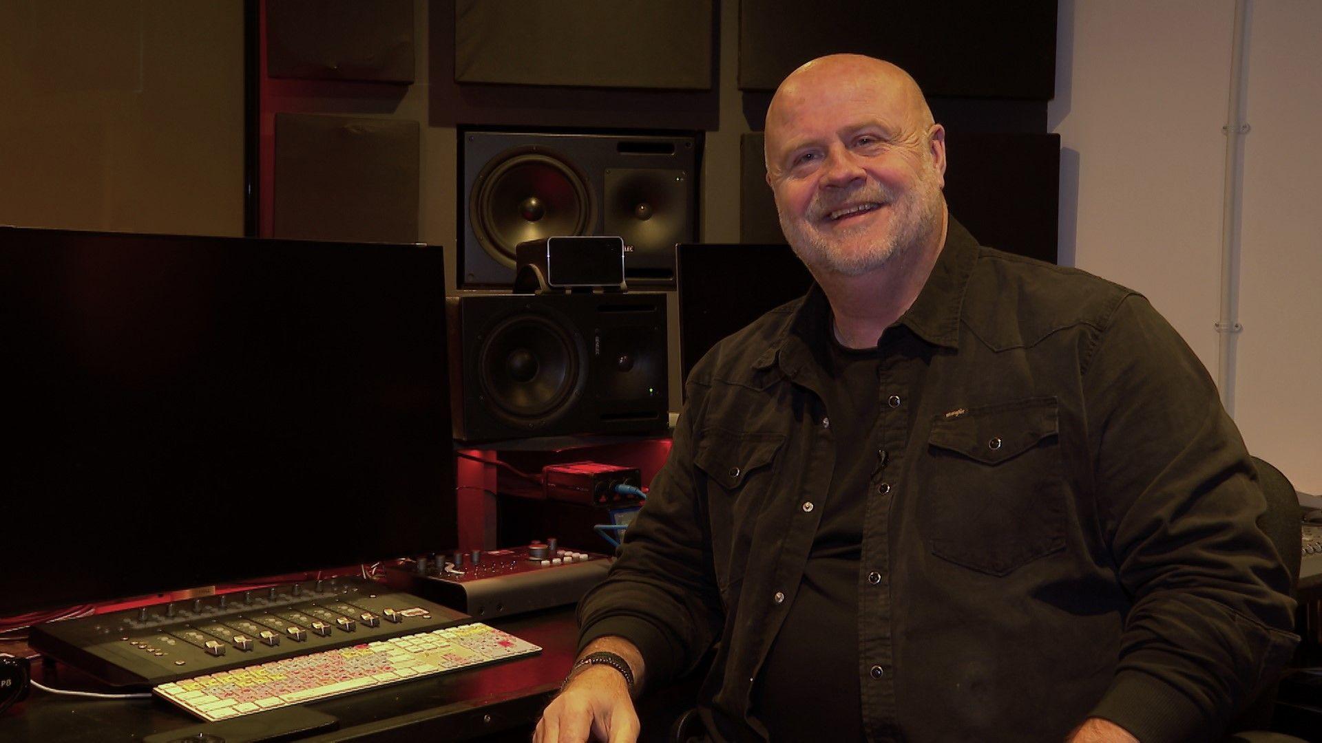 Emyr Afan, a bald man in a dark shirt, smiles at the camera as he sits at a sound mixing desk.