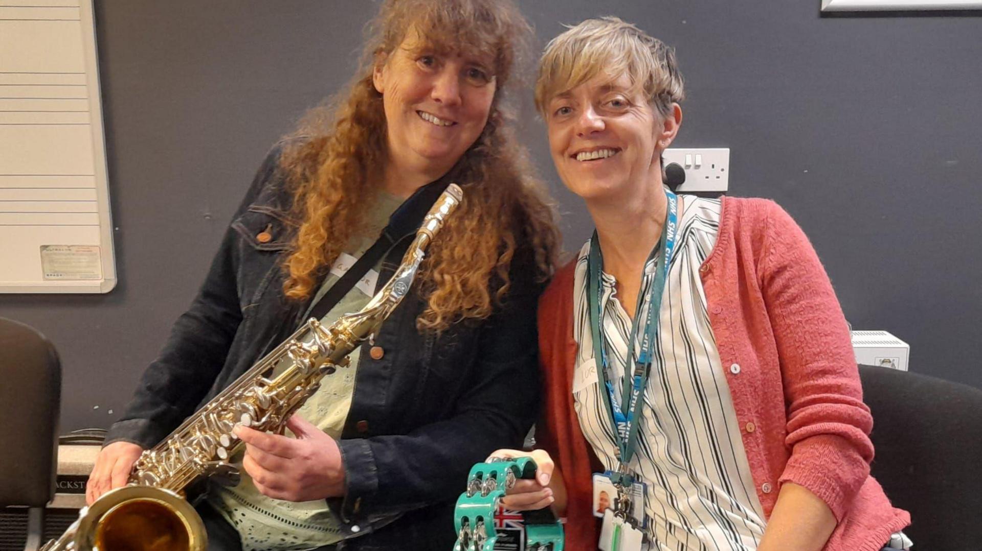 Gaynor has long curly red hair with a fringe, styled in a topknot. She is sat, smiling at the camera, wearing a dark denim jacket and a pale green t shirt underneath and holds a saxophone. Lorna Farmer sits to her right, she is also smiling at the camera. Lorna has short blonde hair with a long fringe framing her face, she wears a blue striped blouse with a coral pink cardigan and has a blue NHS lanyard around her neck and holds a turquoise coloured tambourine.