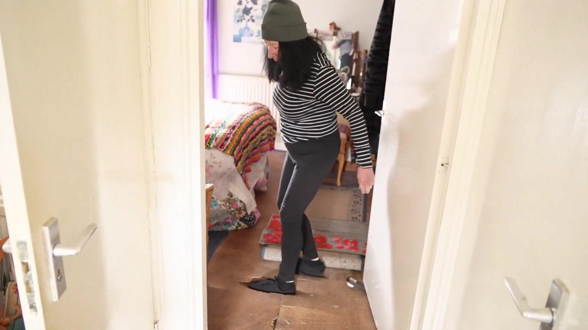 Sharon Lawson, a woman with long black hair wearing a grey beanie hat and black and white striped top, shows where her foot broke through the wooden floor in her bedroom