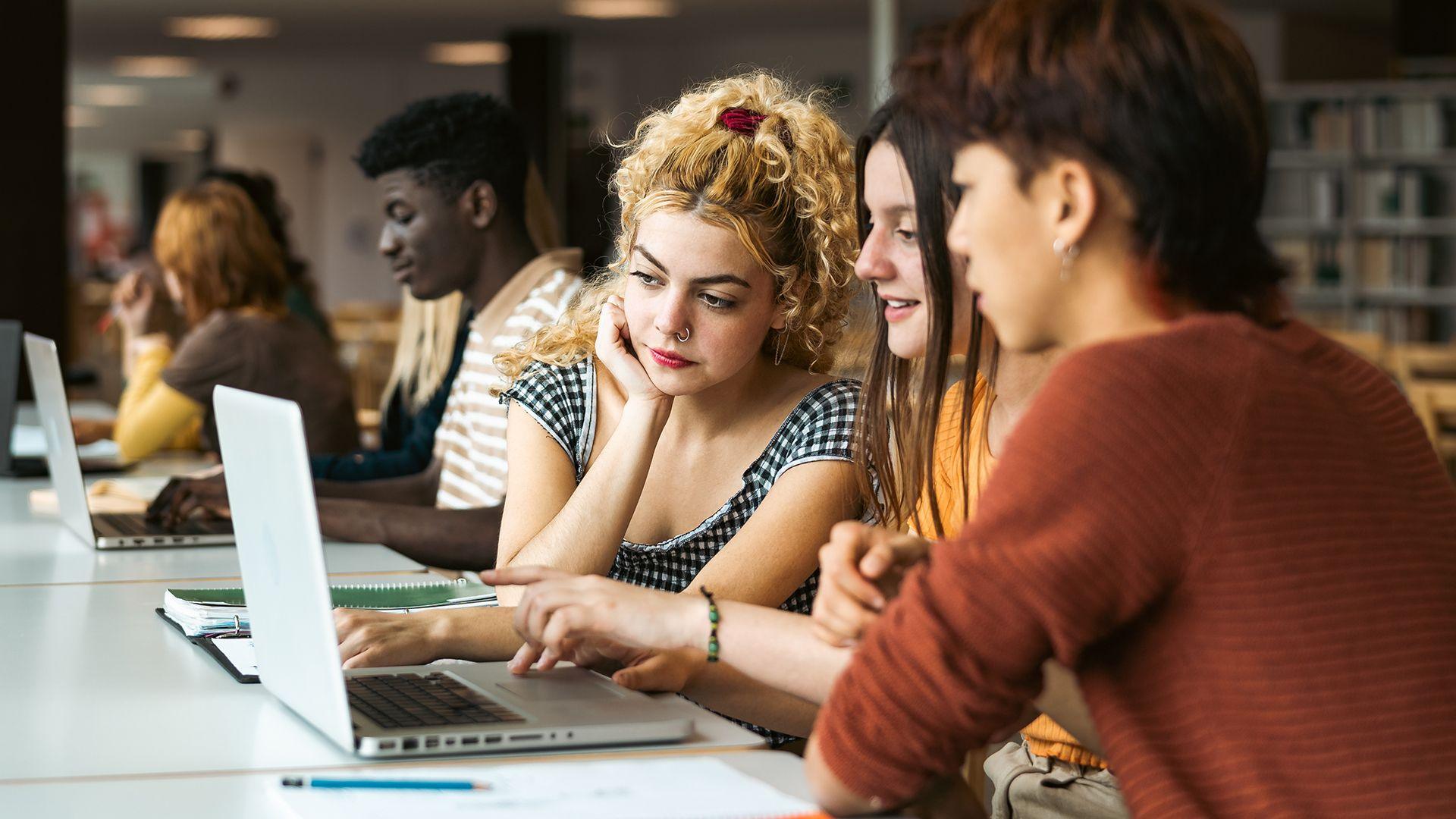 Stock photo shows three students looking at a laptop in a university library with other students and book shelves in the background.