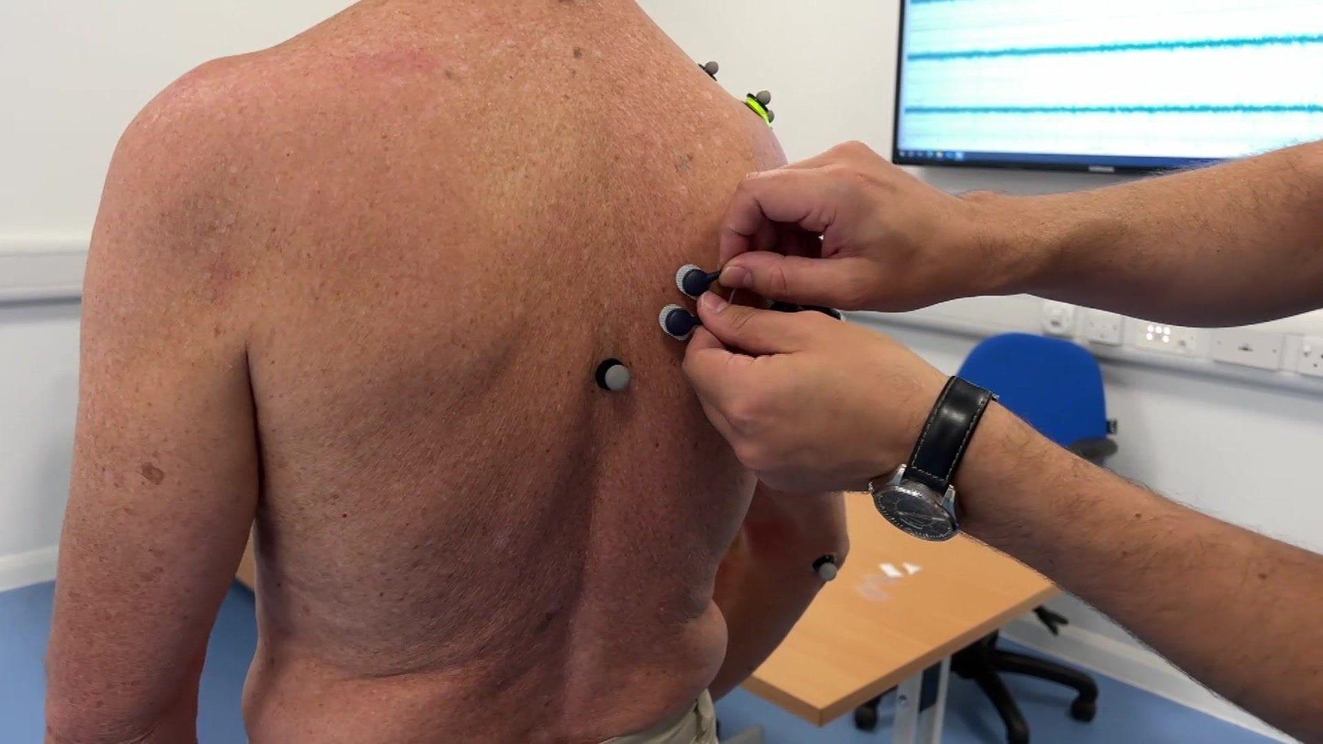 Paul Bradley standing topless while small round grey sensors are attached to the top right of his back