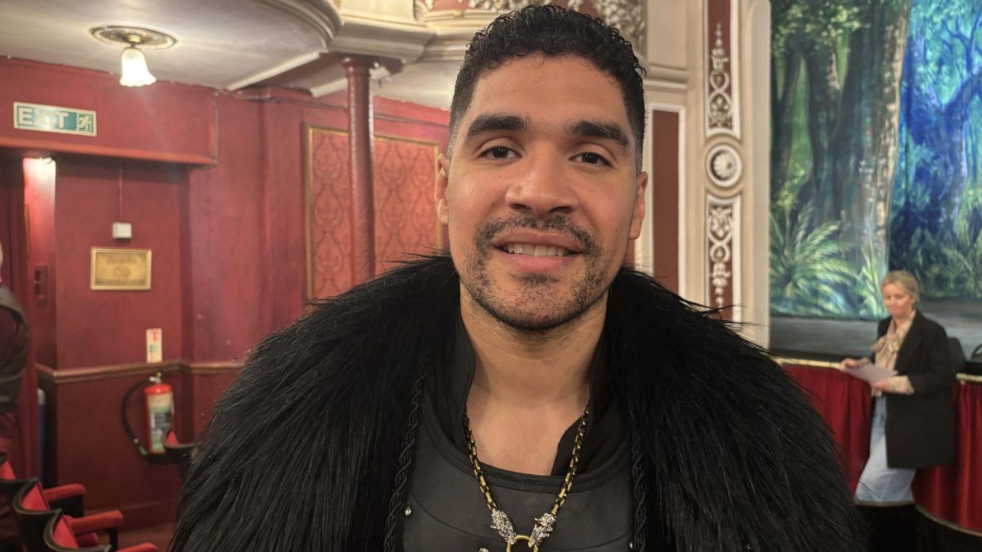 Head and shoulders shot of Louis Smith. He has dark brown hair and a goatee beard and is wearing a fluffy black jacket and chain and smiling at the camera.