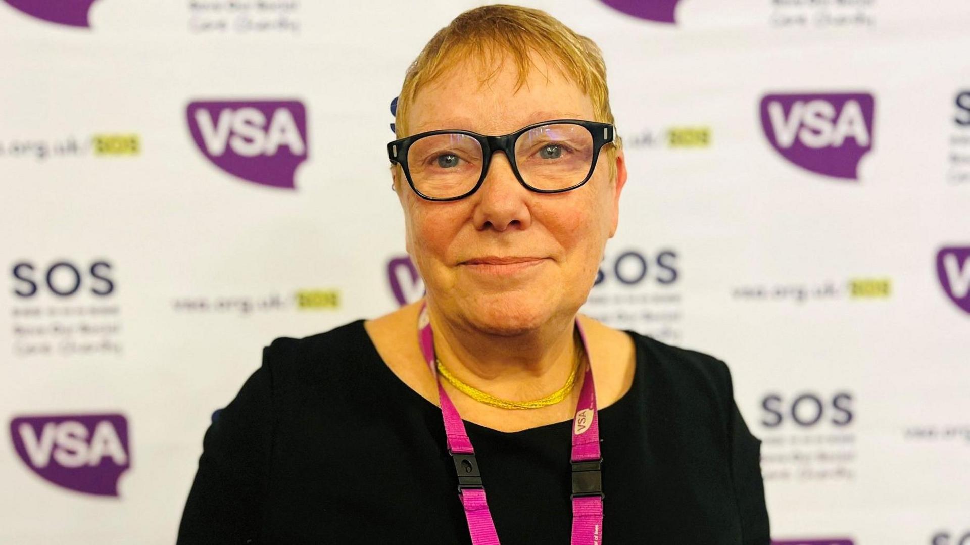 Sue Freeth - she has short hair and glasses with black frames is standing in the middle of the shot and looking into the camera. She is wearing black and has a lanyard on her neck with a charity's logo. The promotional banner with the charity's logo is in the background, that is blurred.
