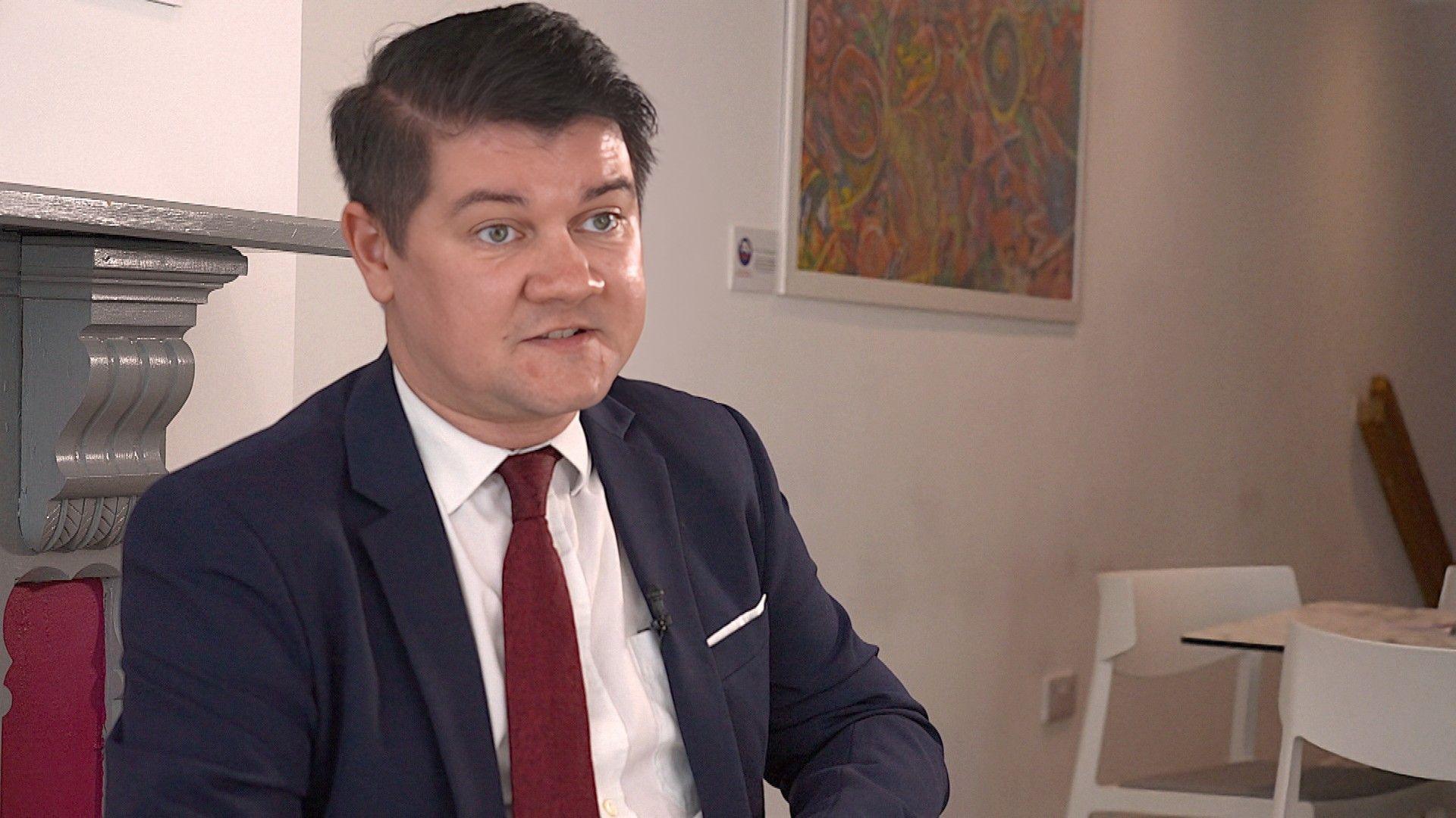 Sam Mezec, , Jersey's Housing Minister, wearing a navy suit, white shift and dark red tie, being interviewed in an office.