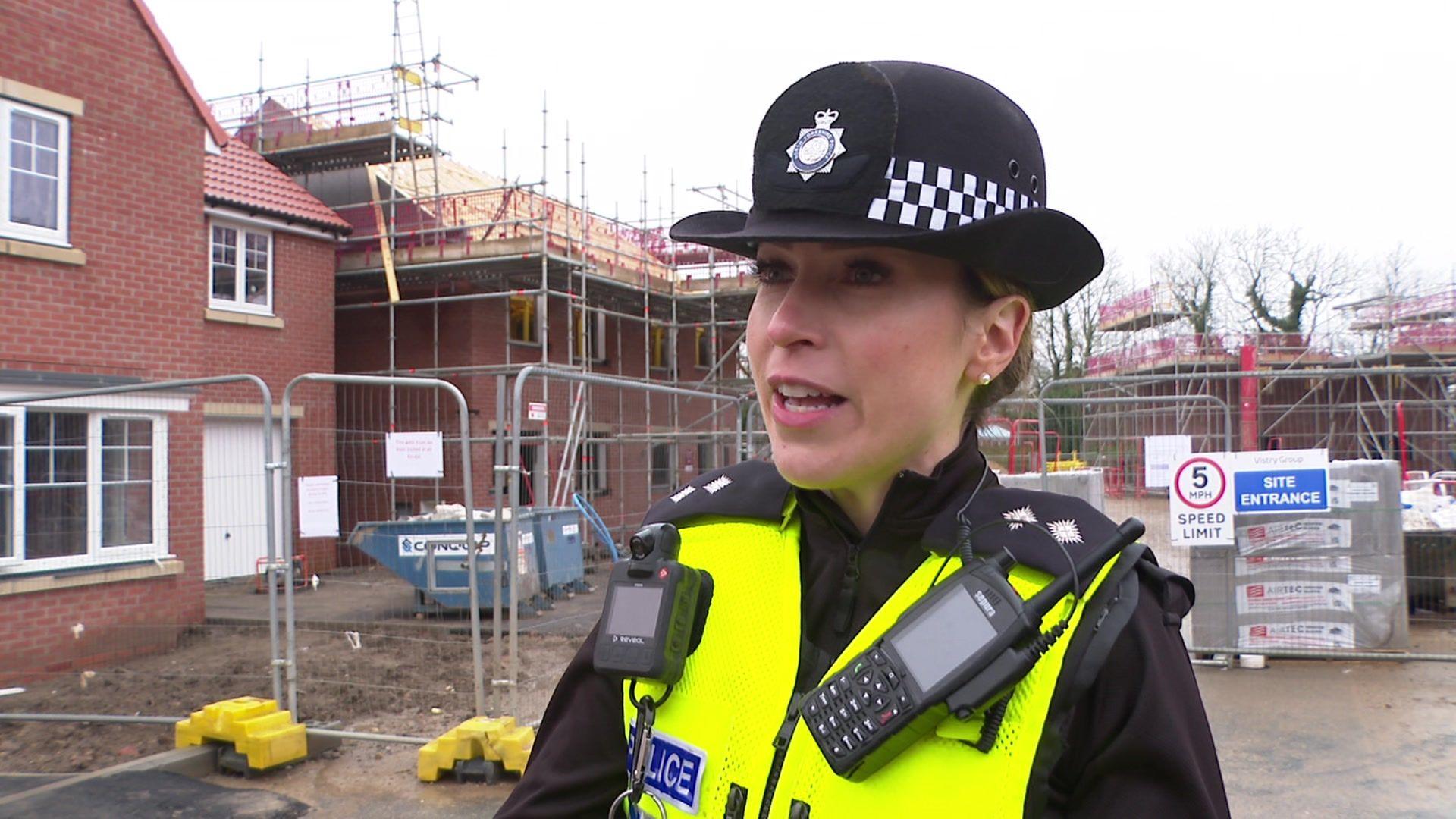 Inspector Holly Nicholls, wearing a high-vis police jacket and police hat standing in front of a red-brick house with scaffolding and fencing around it. 