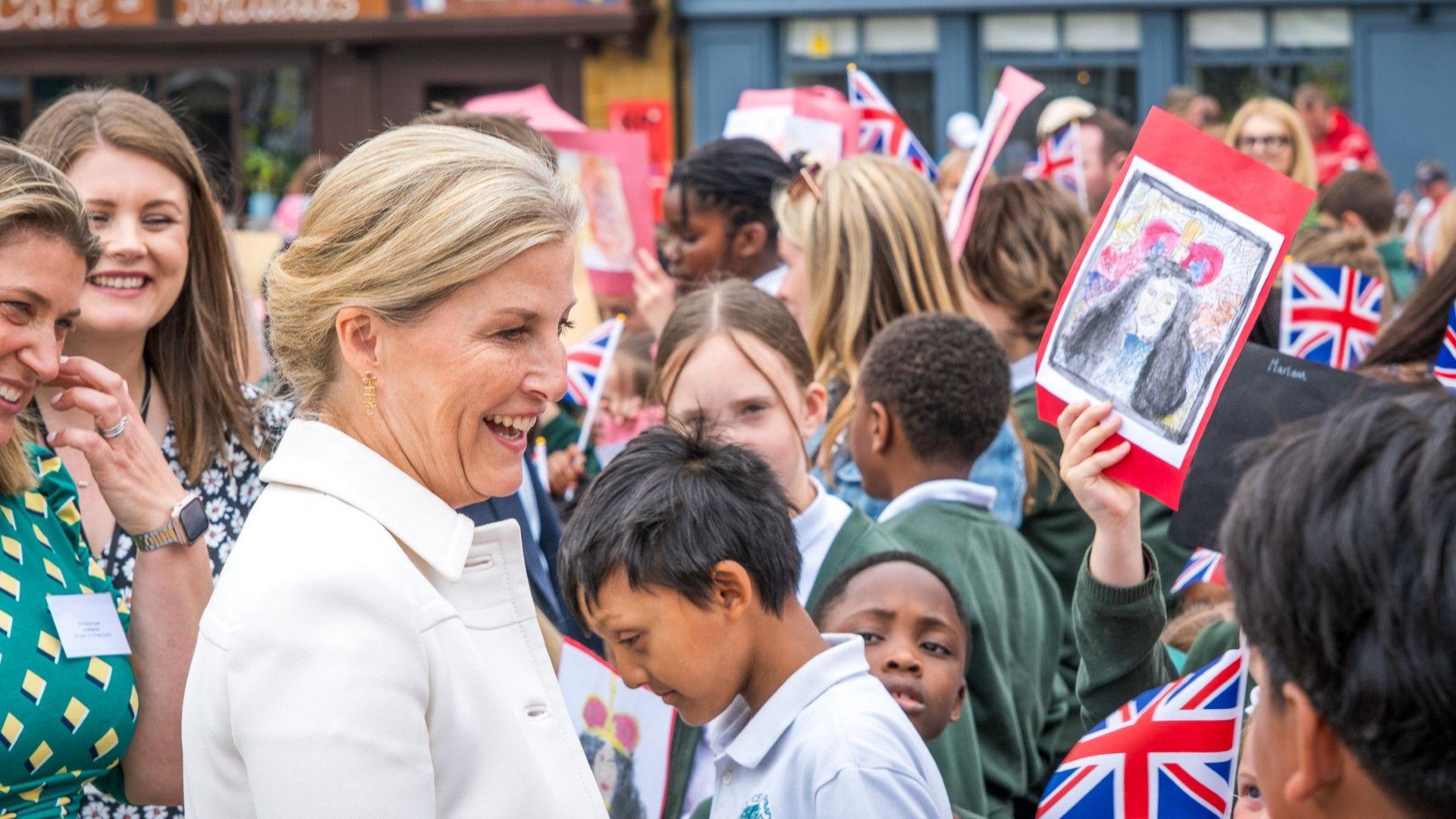 Sophie, Duchess of Edinburgh, in Northampton, talking to school children, they are crowding round her holding flags, pieces of artwork and many are wearing green jumpers. She is wearing a cream jacket and has her blonde hair tied up. Two women are behind her and smiling. 