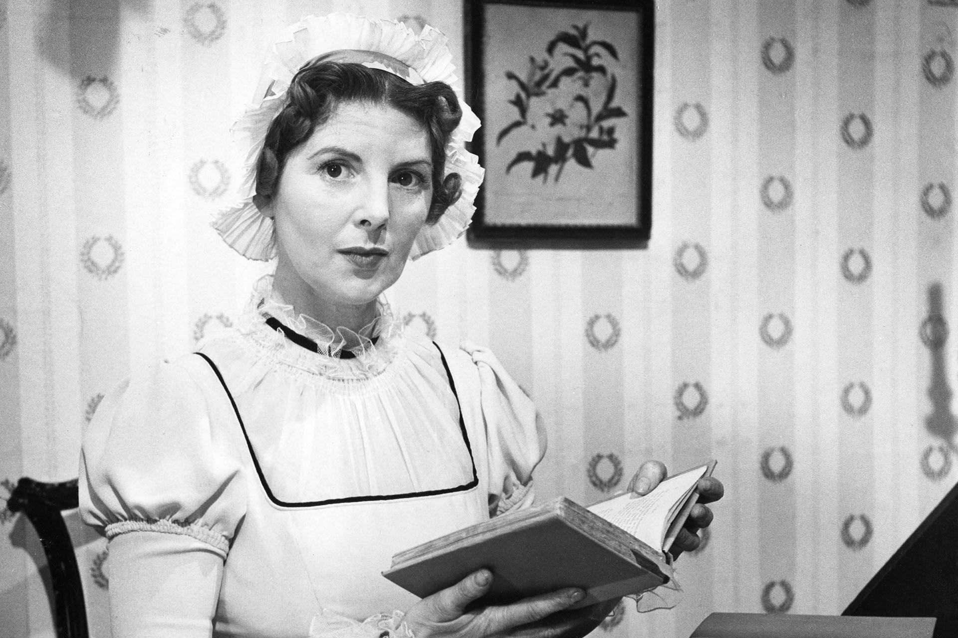 a black and white still of thea holmes sat down and dressed in a high necked white dress and matching bonnet. she holds an open book in her hands