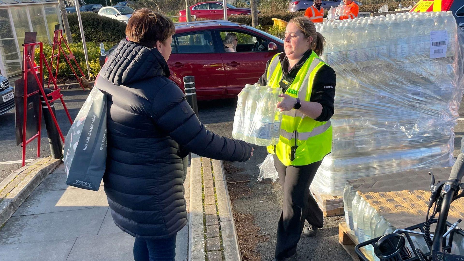 A woman holding plastic wrapped bottles of water is passing it to a second woman. The first woman is wearing a yellow jacket.