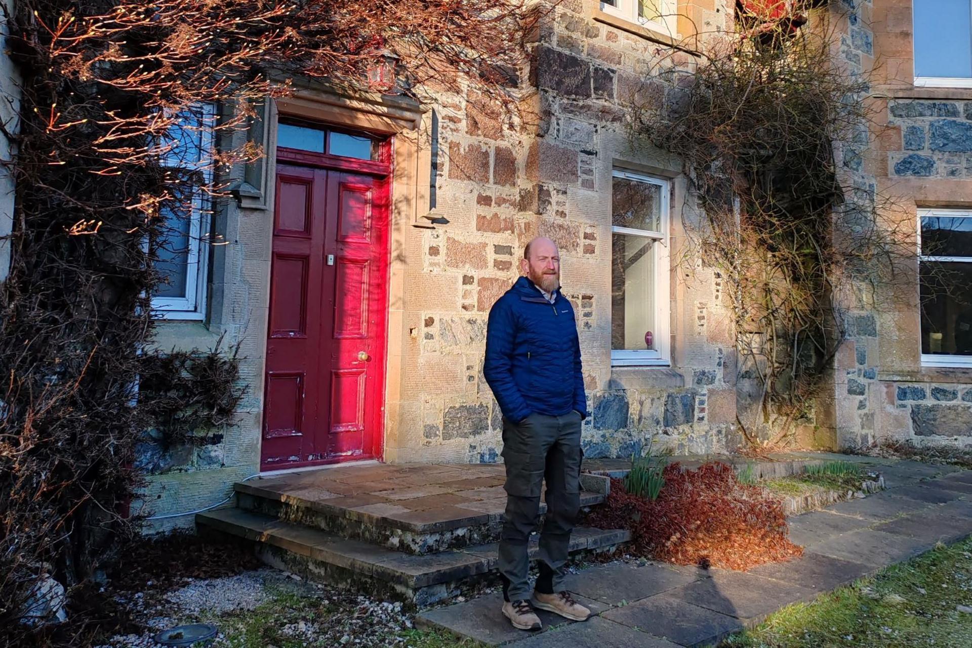 Lewis is standing outside a red door of  Glencanisp Lodge, a former Victorian hunting lodge.