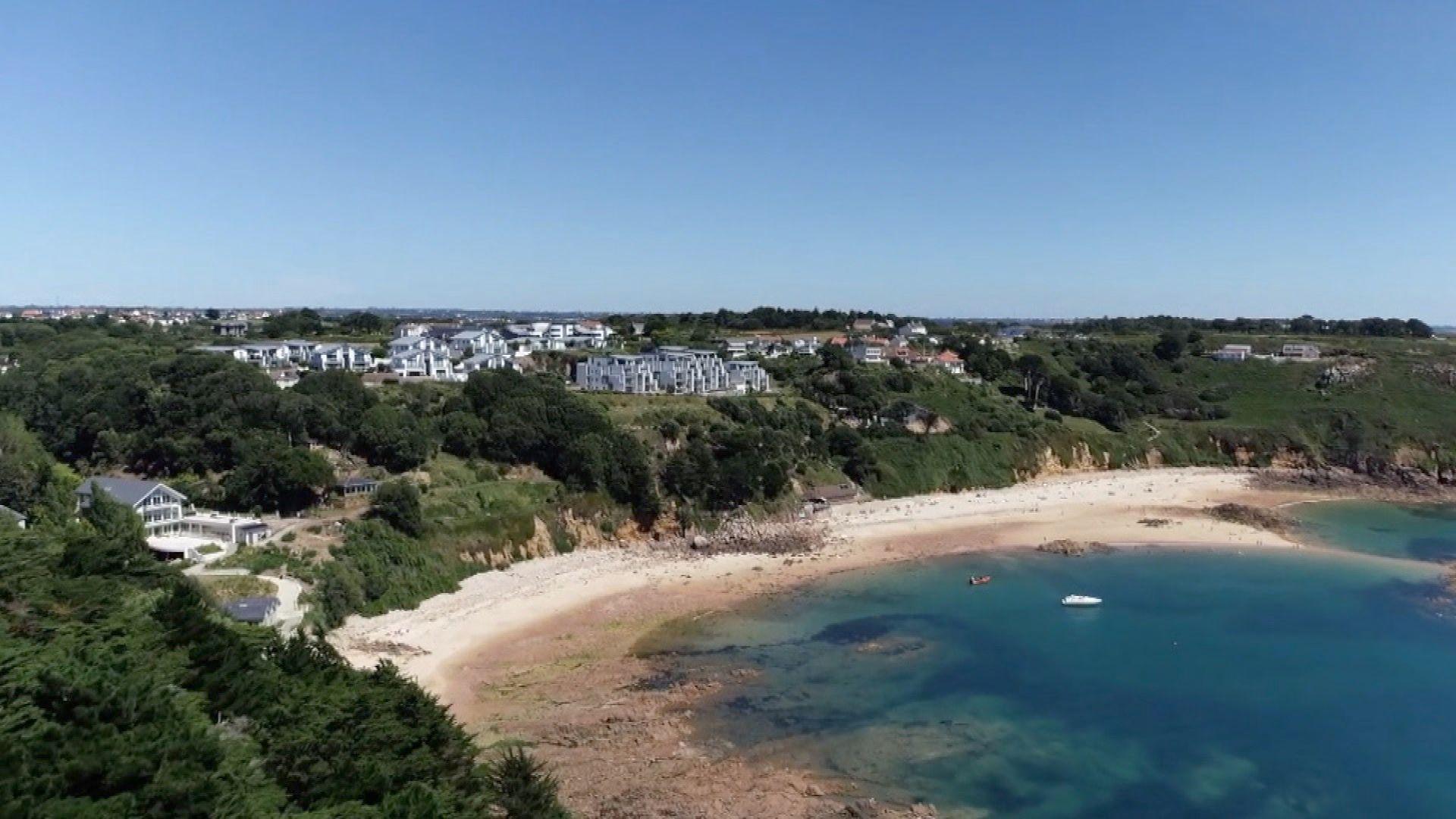 A drone shot looking down at Portelet Bay. There is land, with houses, on the left and a sandy beach to the right. The sea is blue and is clear. The sky is blue.
