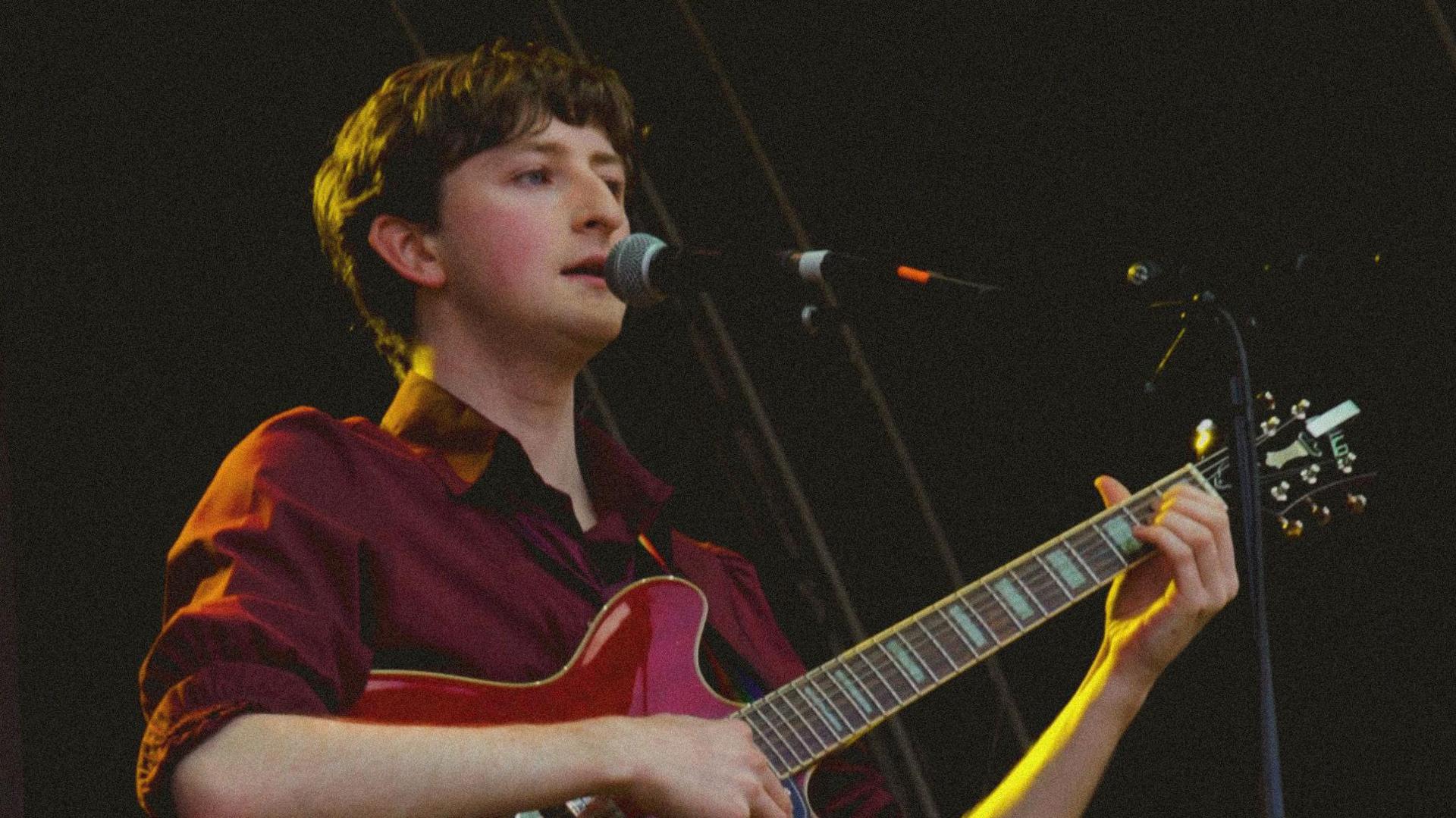 A man stands on stage singing into a microphone. He is playing a pink electric guitar and wearing a burgundy shirt.